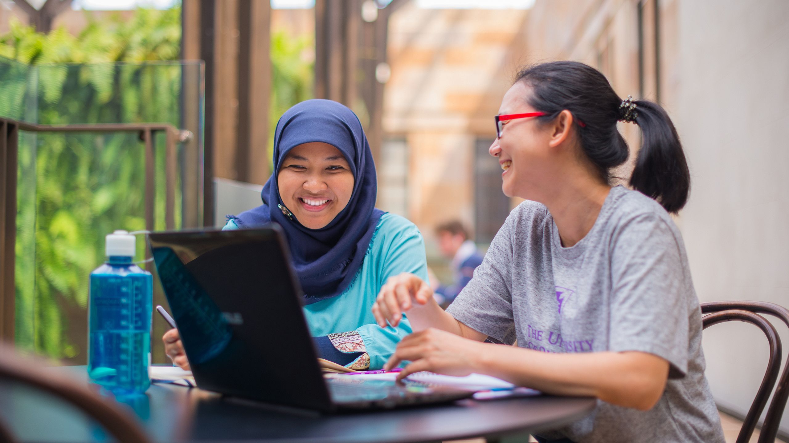 Two smiling women sitting at a table with a laptop in front of them.