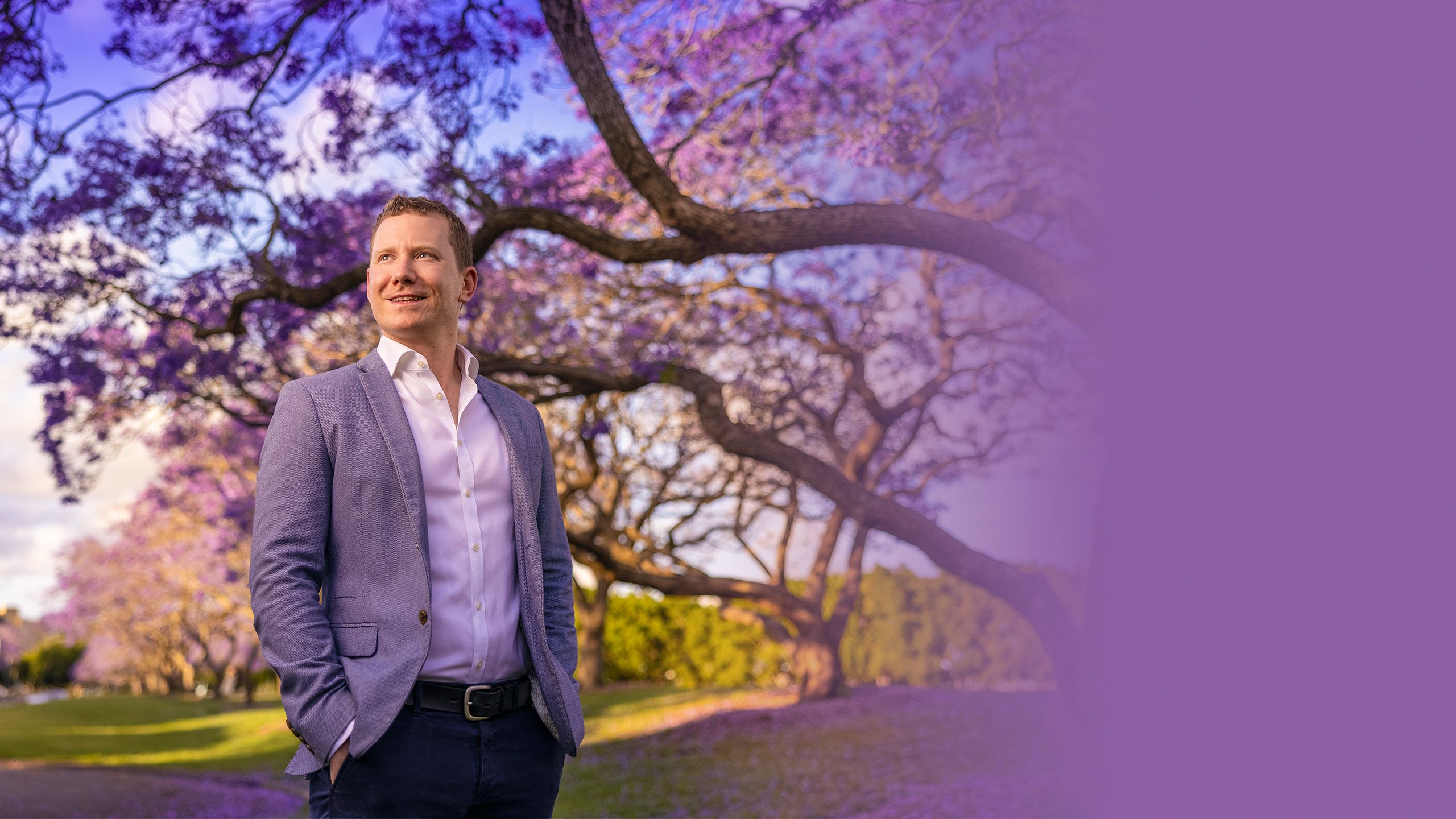 Image of Daniel Wessels a smiling young man with blonde hair wearing a purple suit jacket, standing under a blooming purple jacaranda tree with fallen jacaranda flowers scattered on the ground