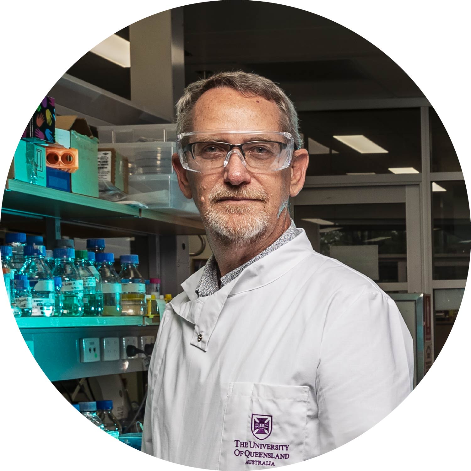 A man in a white UQ labcoat and safety goggles looks seriously into the camera. He is in a lab, with bottles of liquid behind him. He has grey hair and a beard.