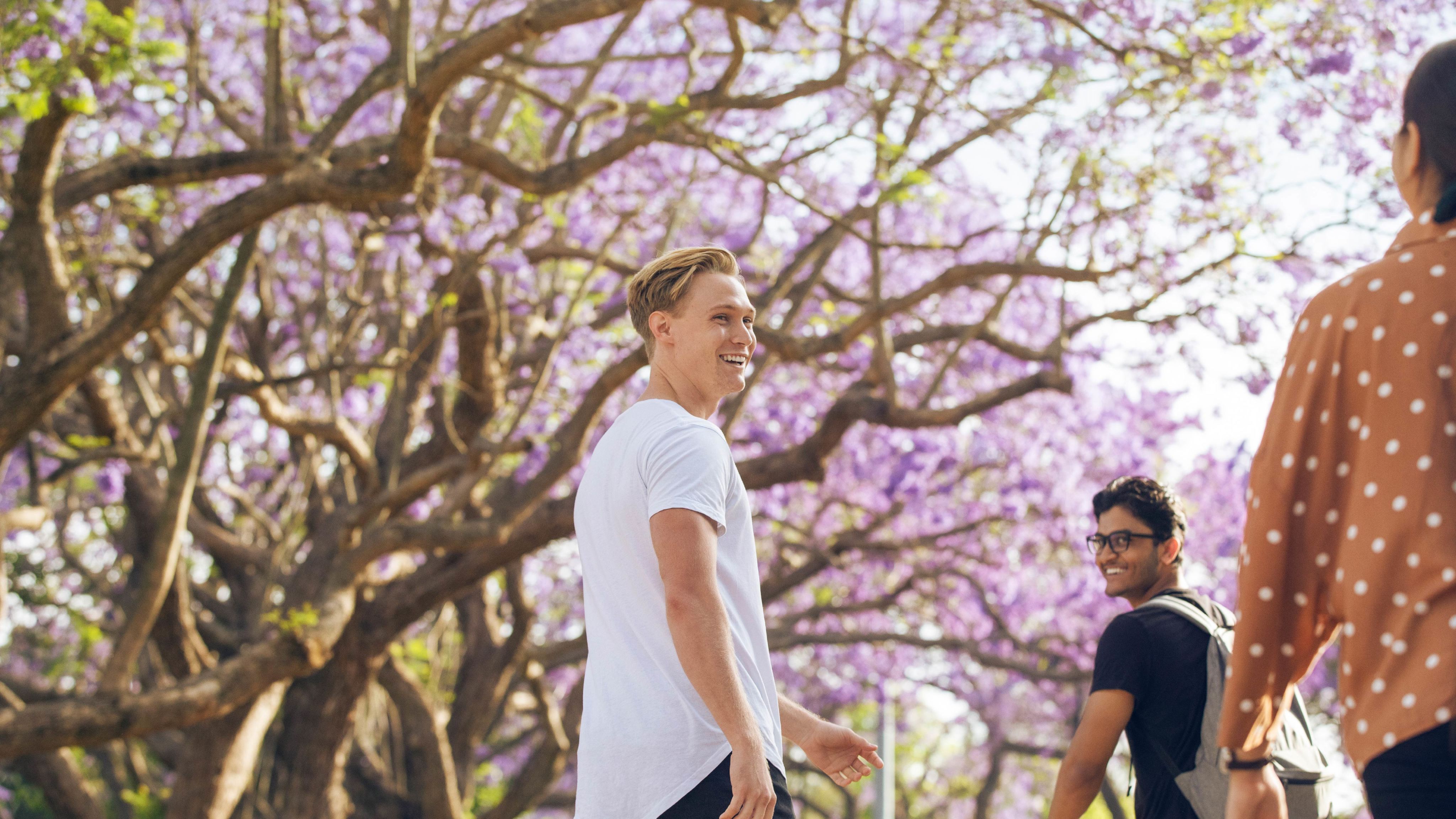A young man with blonde hair looks behind himself while walking beneath a blooming jacaranda tree.