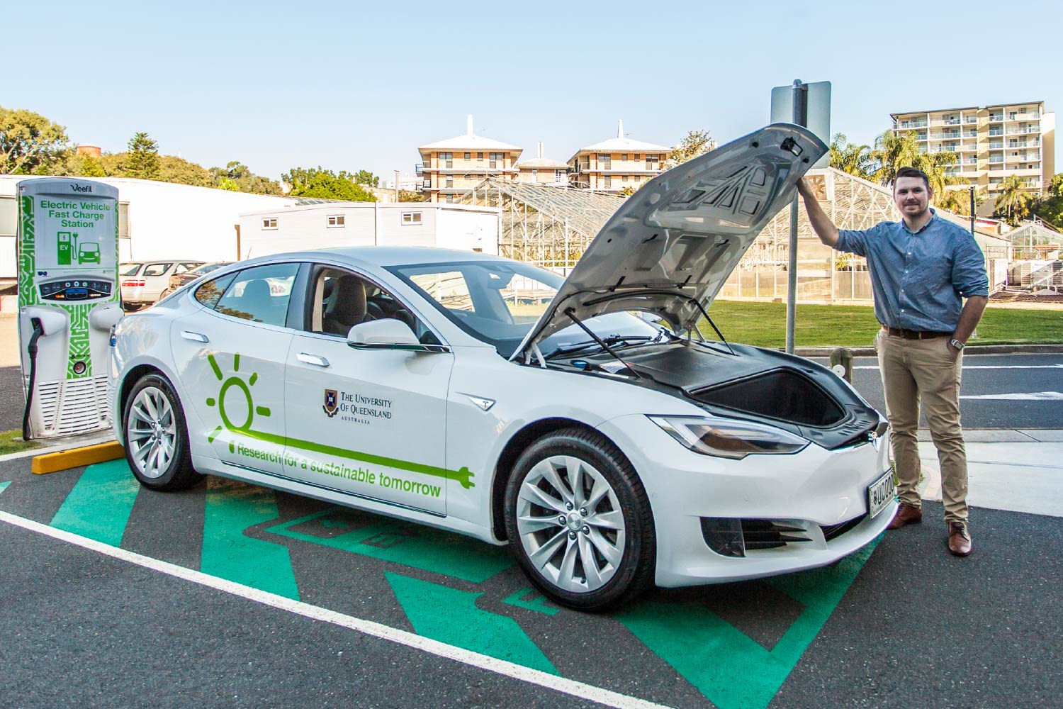 A man holds open the bonnet of an electric UQ vehicle that is charging in a charging spot. He is on the UQ campus.