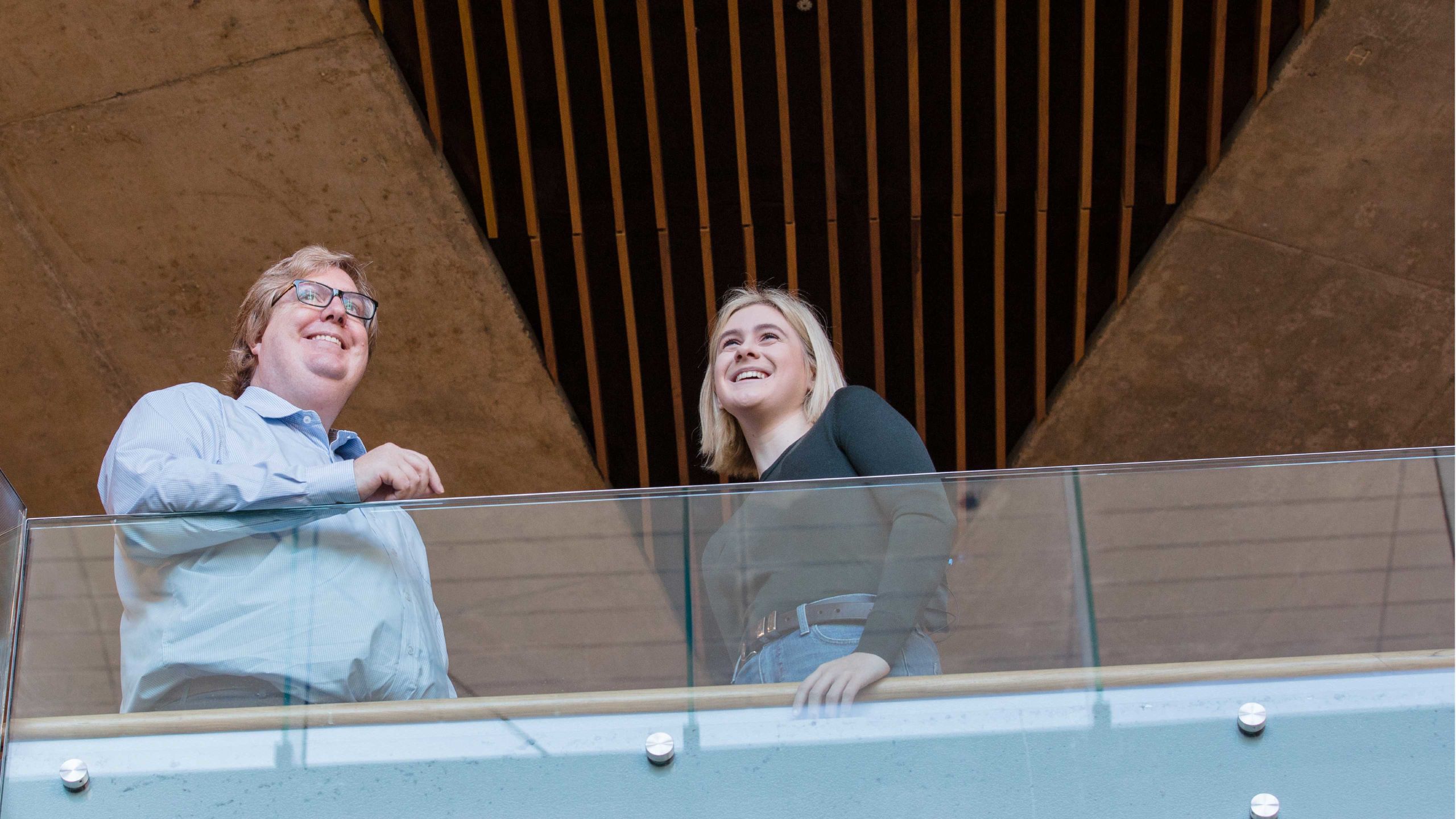 A university professor and a student smile as they looks out over a modern glass balcony.
