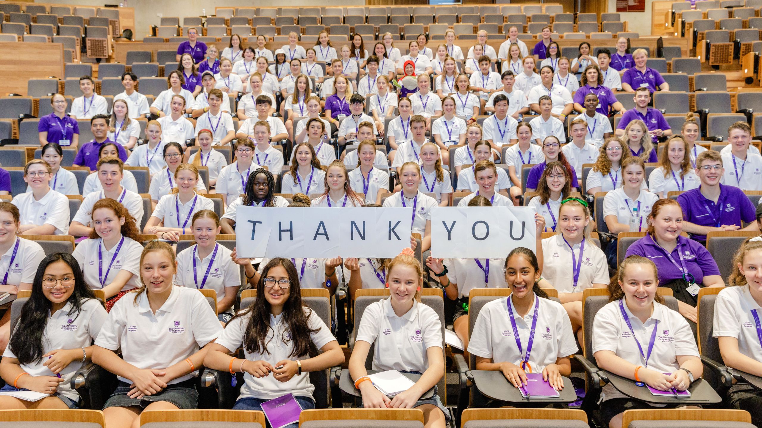 A diverse group of 50 or more students from a range of cultural backgrounds sit in a tiered lecture hall wearing purple lanyards. The middle five hold a big paper sign reading 'thank you'. Everyone is smiling.
