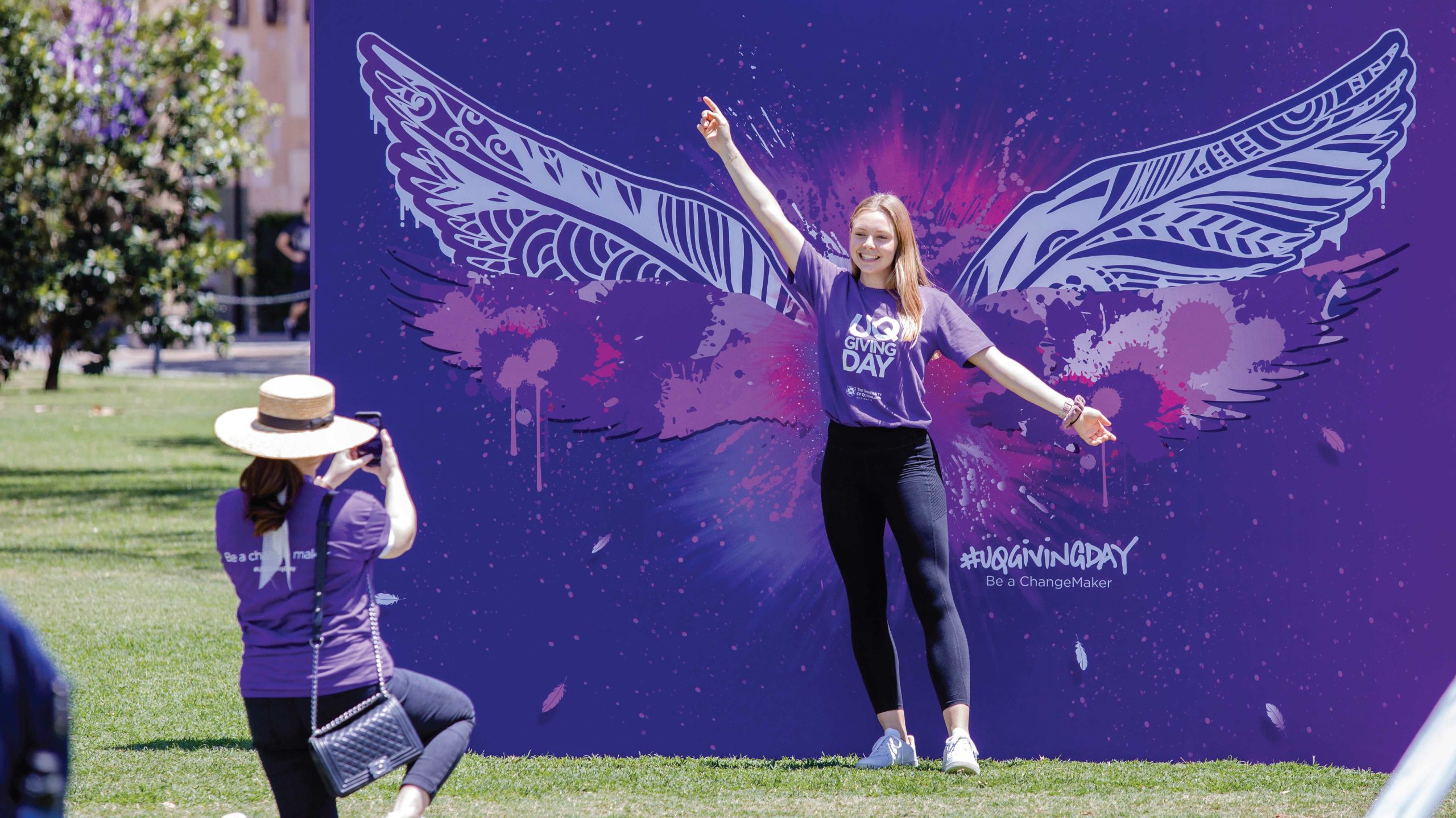 One woman takes a photo of another woman posing enthusiastically in front of a mural with angel wings.