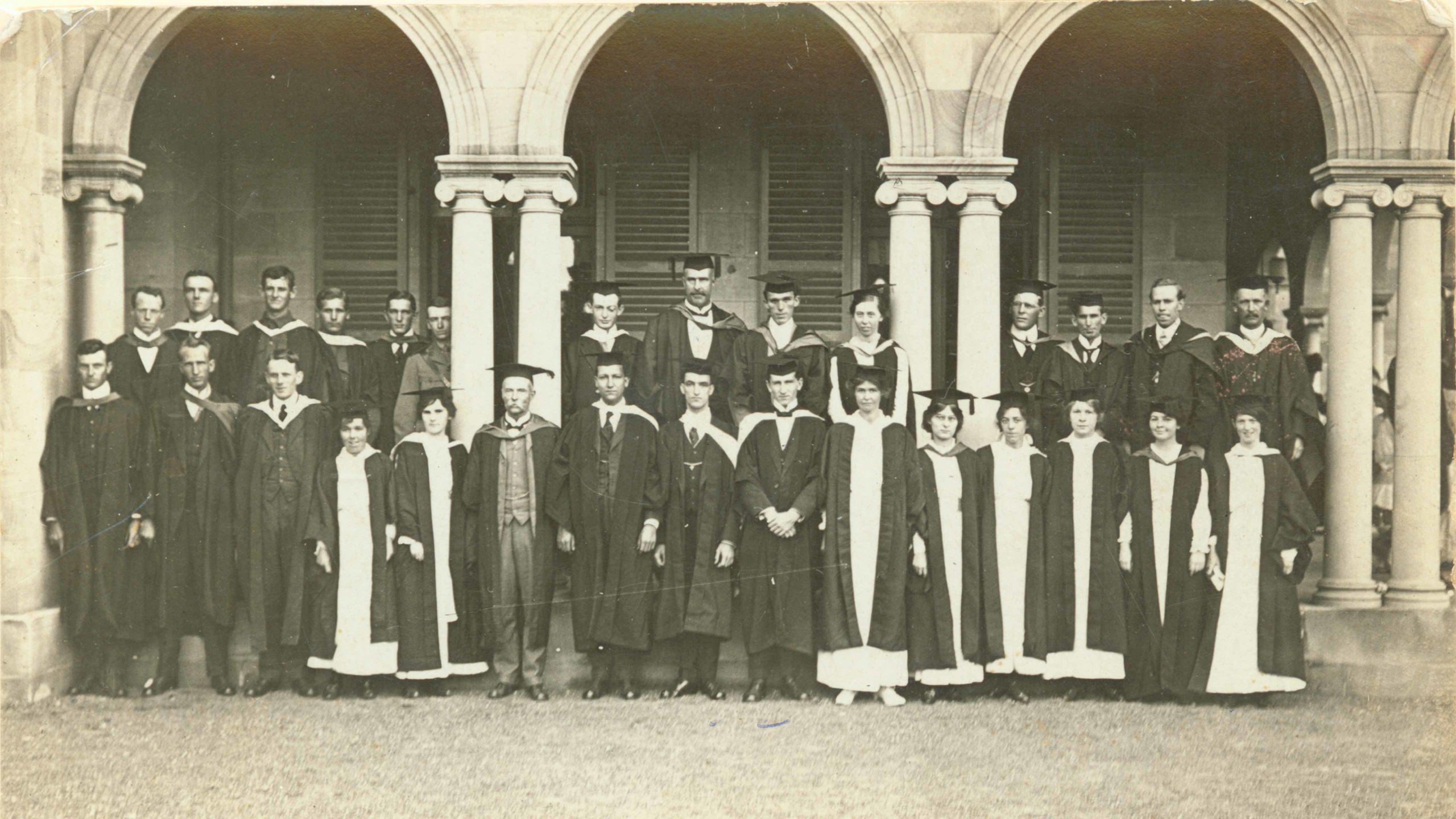 Around twenty men and women in academic dress stand in front of a grand building with pillars and archways, posed in two rows.