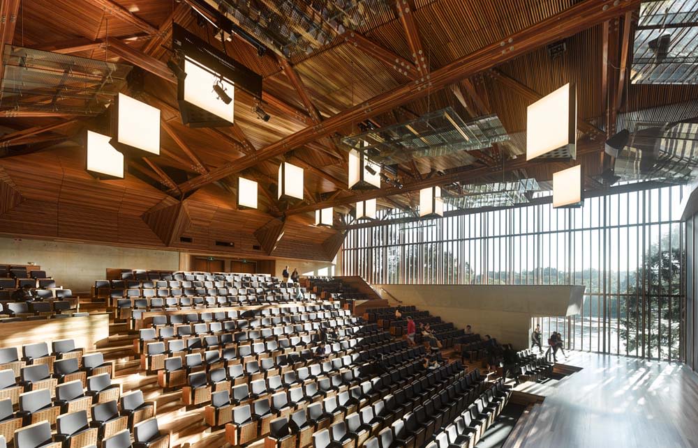 A large, modern lecture theatre, architecturally designed, featuring raked timber ceilings and floor to ceiling windows with an outlook over a lake.