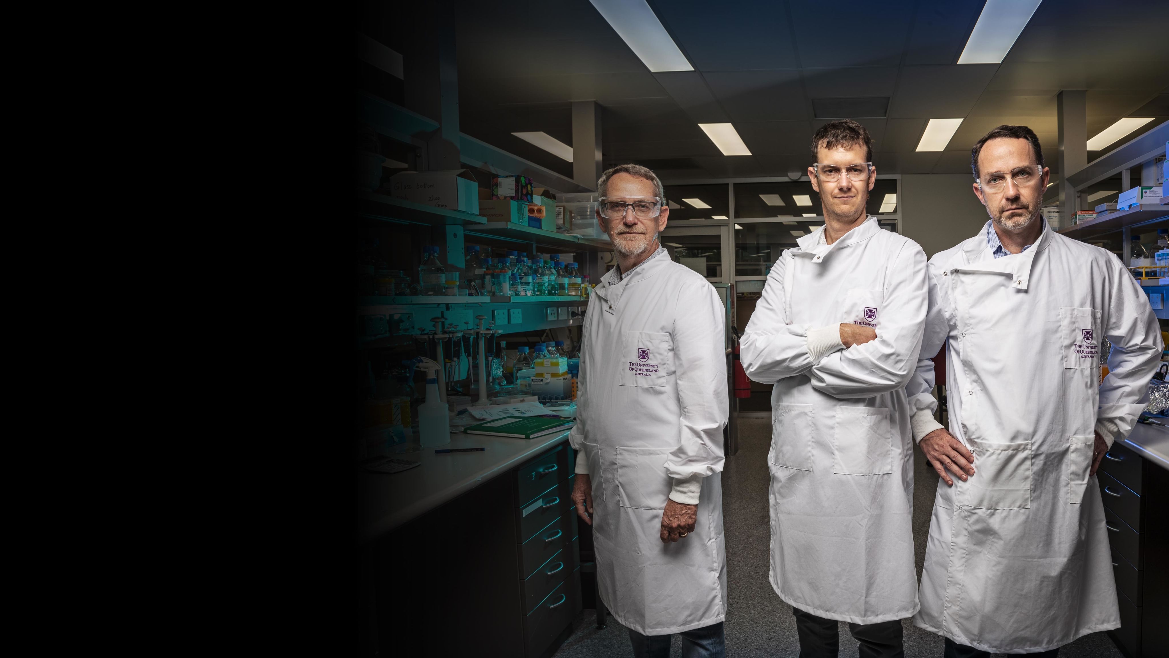 Three medical researchers wearing white coats and safety goggles look directly to camera while standing in a lab setting.