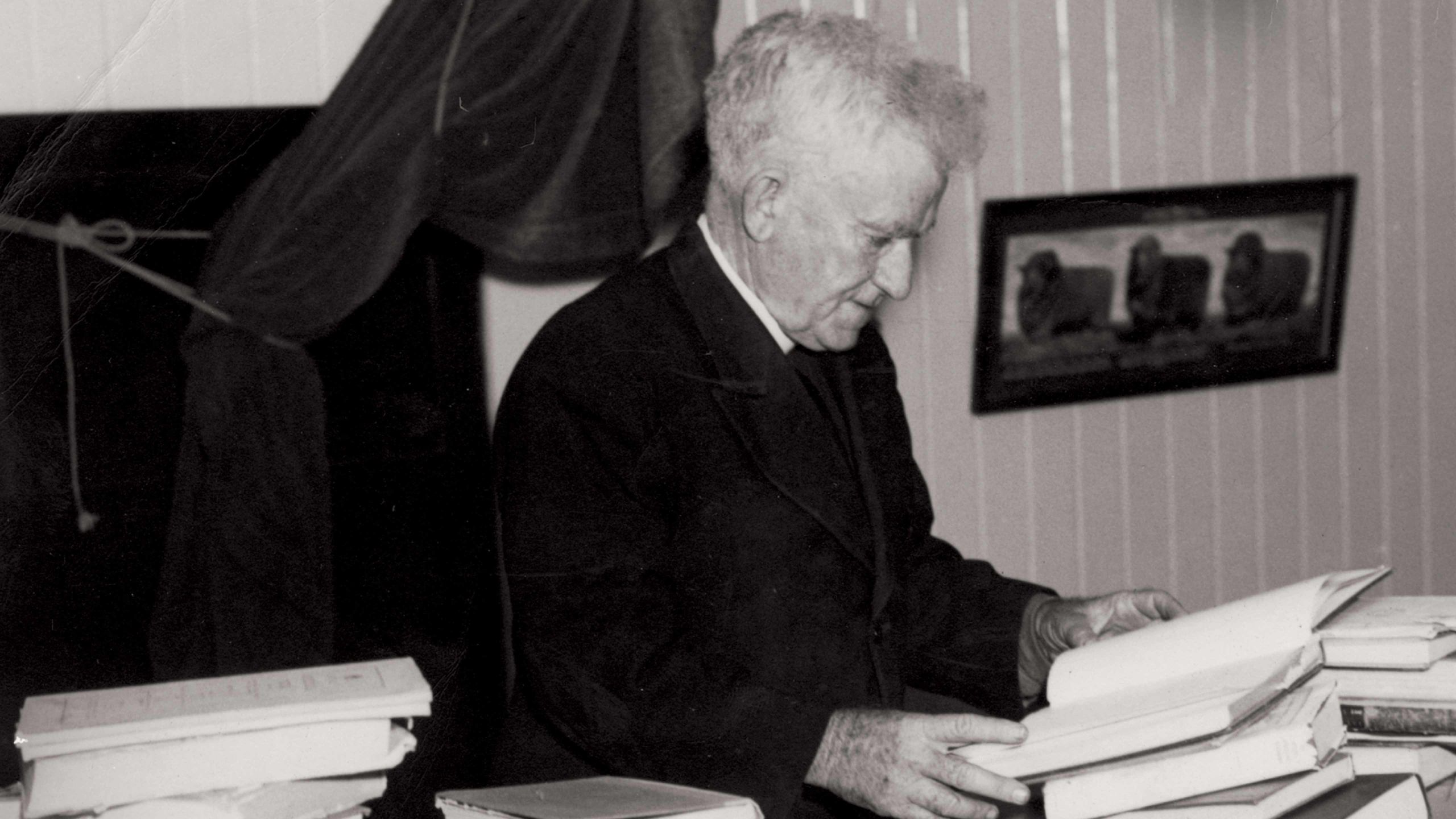 An elderly man in a priest uniform flicks through a book piled amongst others on his desk.