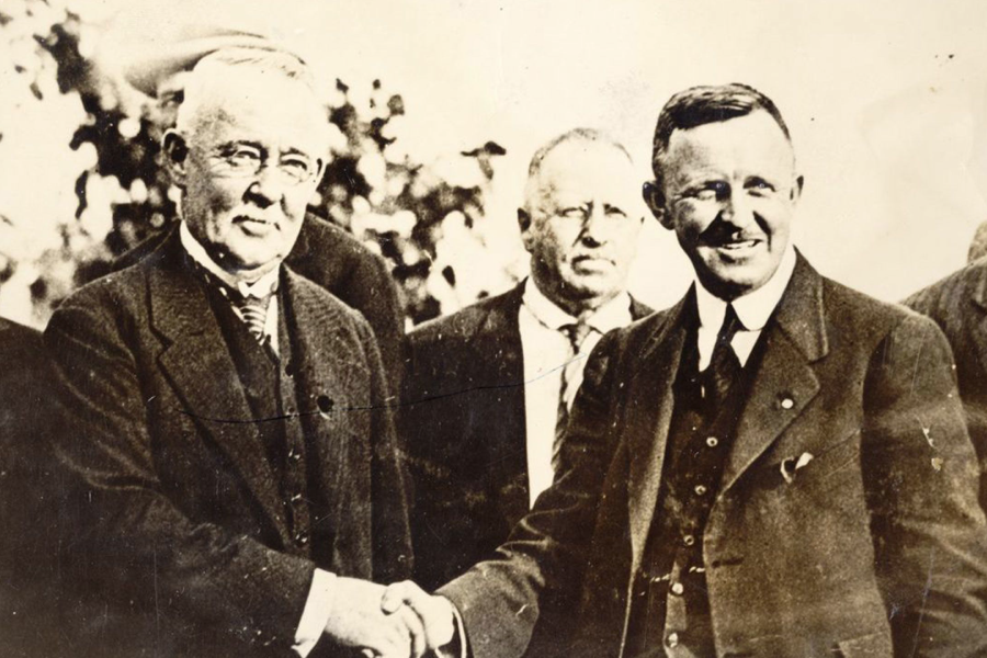 A sepia-toned image shows two men in three-piece suits shaking hands and smiling.