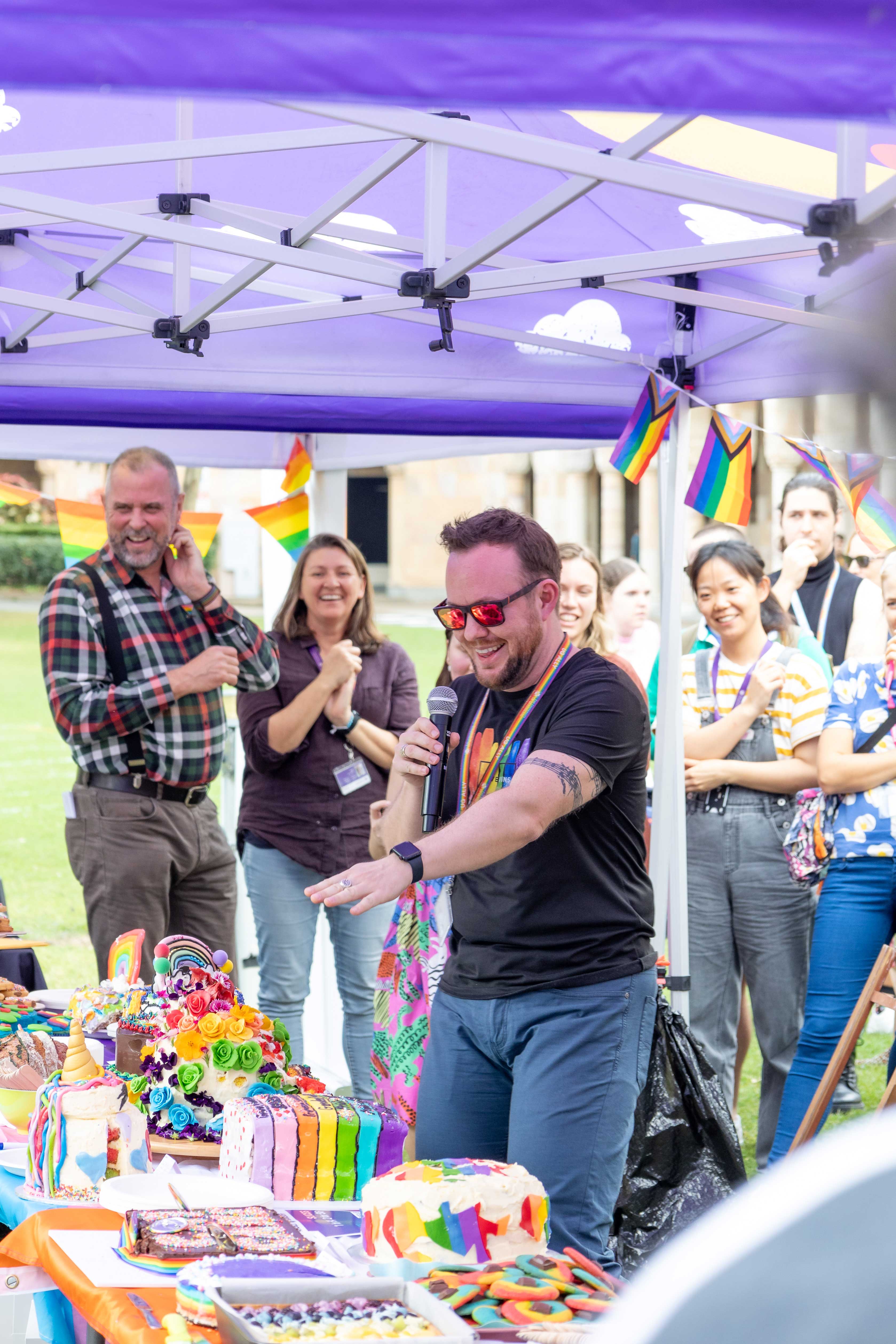 A person presents at the Annual Great Rainbow Bake Off, gesturing to the cakes.