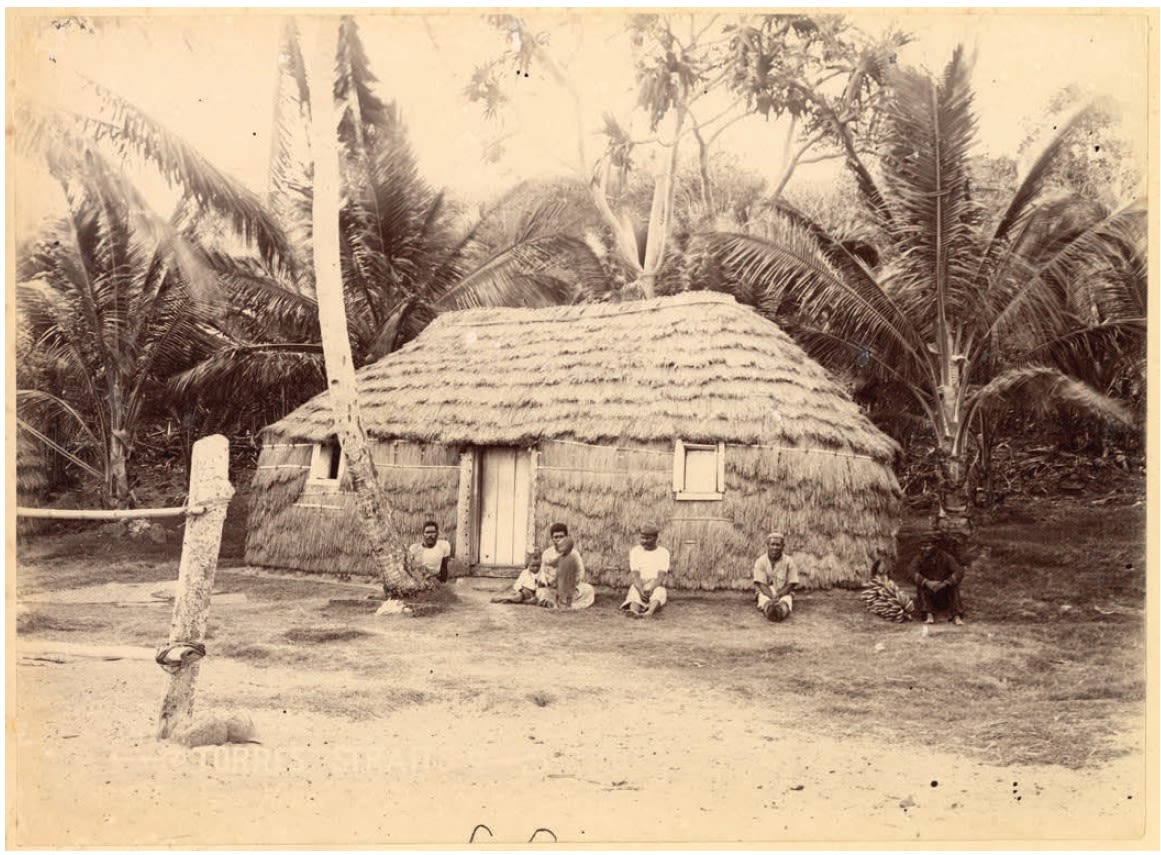 Four people sitting in front of a grass house on a beach in front of palm trees. The photograph is old and is in black and white.