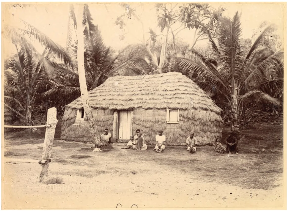 Four people sitting in front of a grass house on a beach in front of palm trees. The photograph is old and is in black and white.