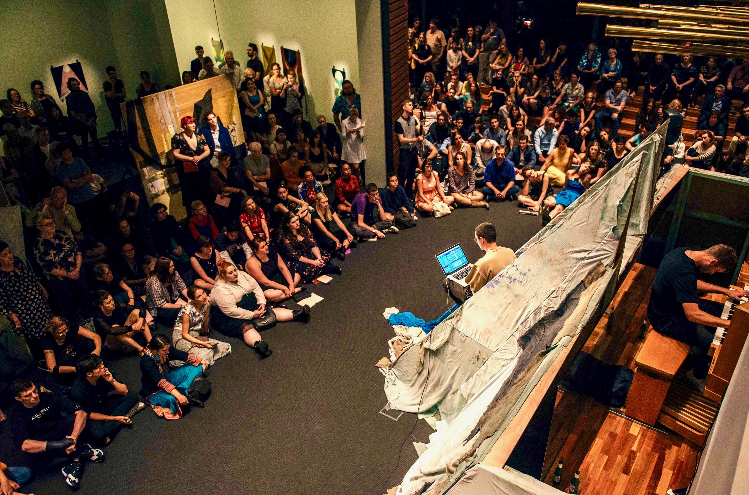 A crowd is gathered for a performance by Clare Milledge with Austin Buckett, Tom Smith, and Luke Pigliacampo of I am an infant: who but I peeps from the unhewn dolmen, arch? I am the tomb: of every hope 2019 at the opening of the Second Sight exhibition at UQ Art Museum..