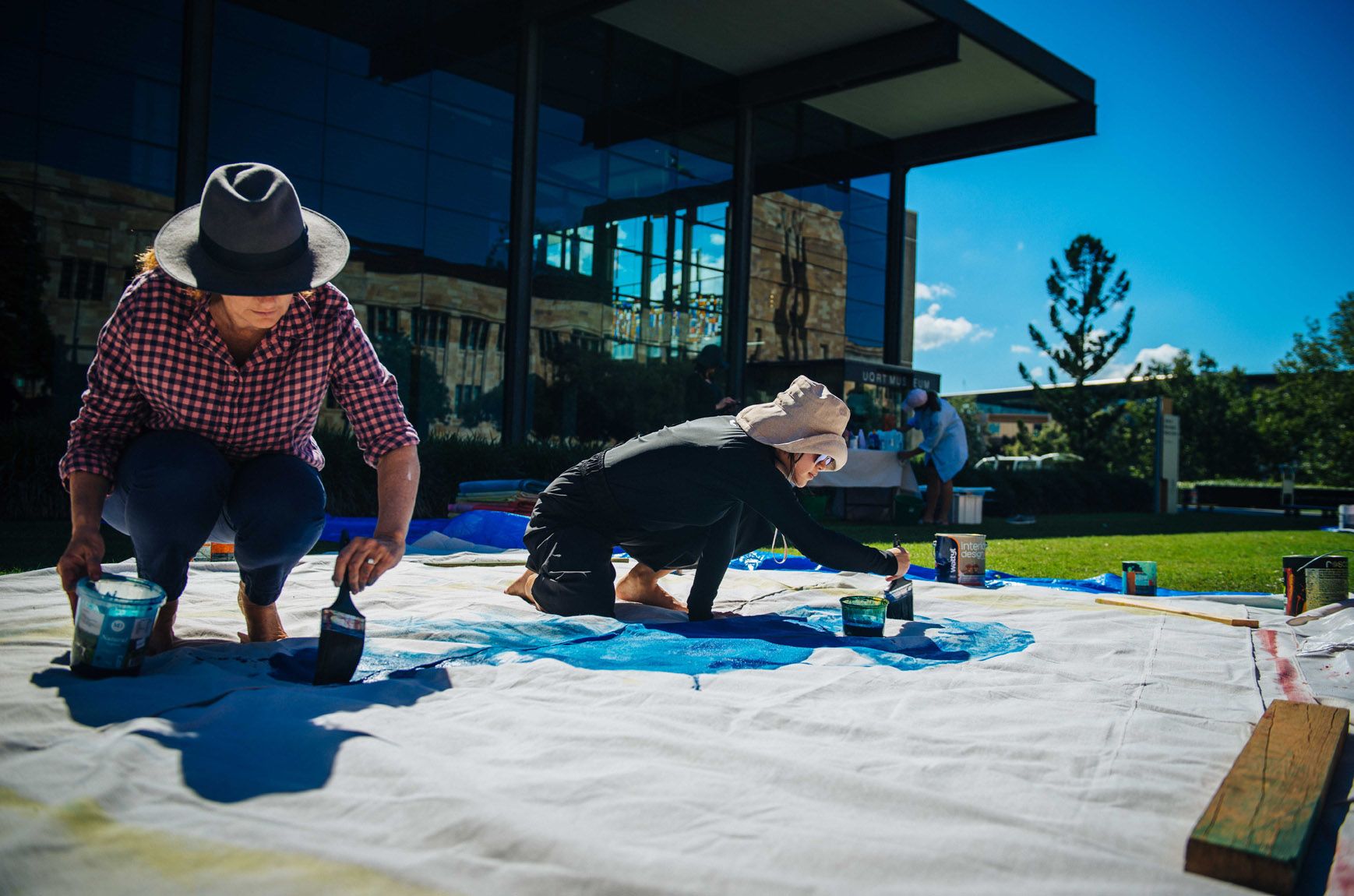 Artist Lara Merrett paints a canvas laying flat on the grass outside UQ Art Museum with the help of a UQ student volunteer on a sunny day.