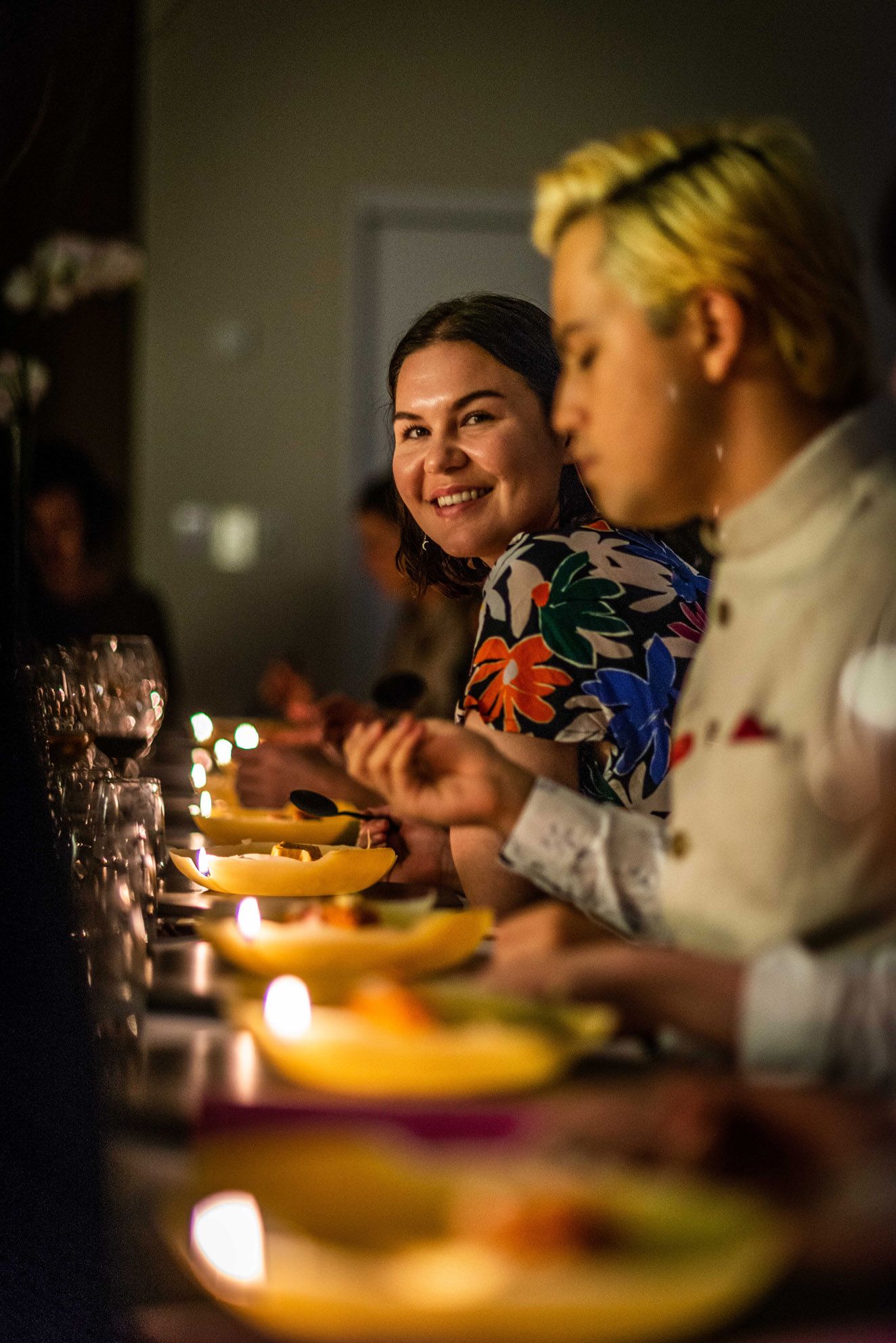 Olivia Trenorden sits smiling in focus alongside another guest at the table set with candlelit plates at the Elizabeth Willing concept dinner.