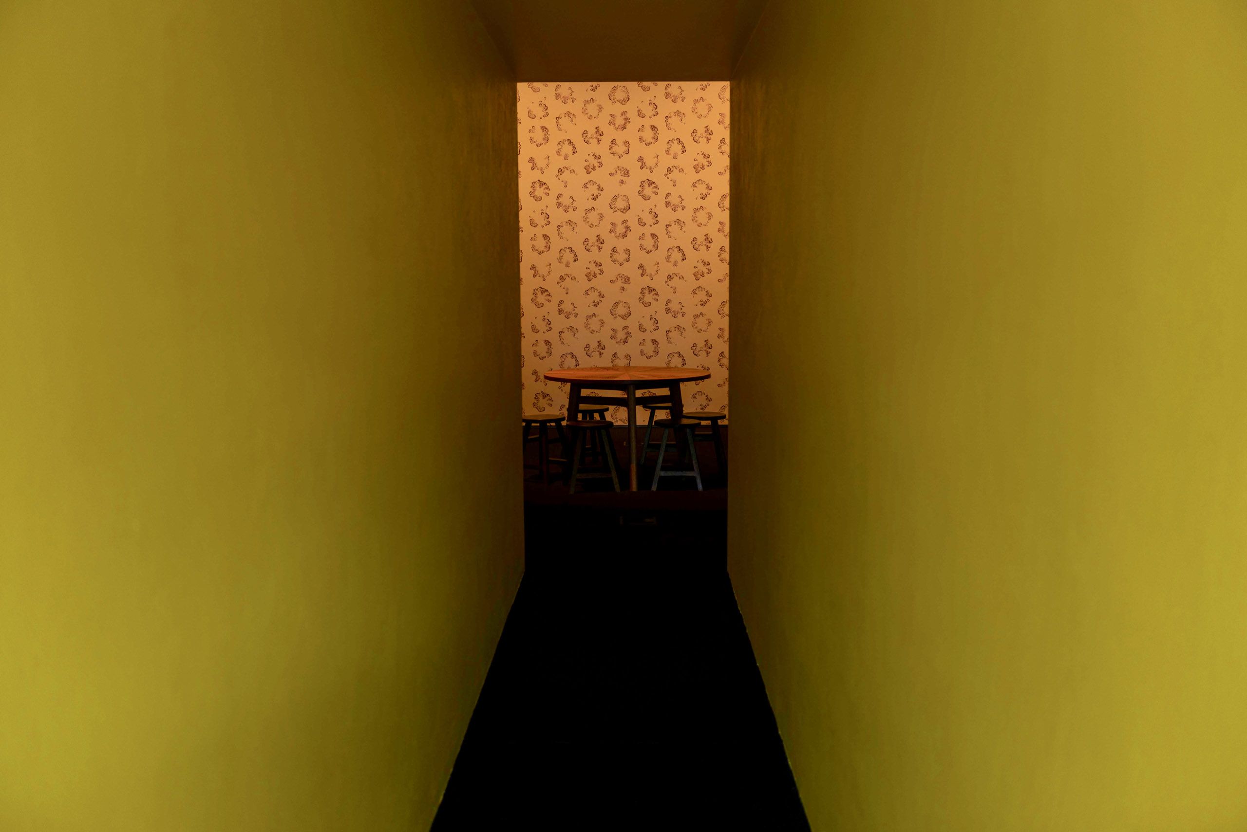 At the end of a bright yellow hallway sits a table against a printed wallpaper backdrop which forms part of Elizabeth Willing's Through the Mother exhibition.