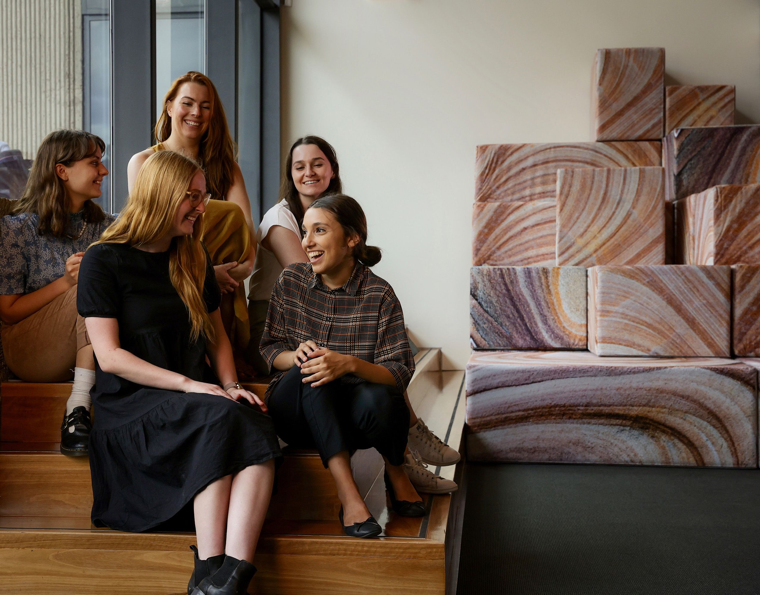Five young women who were UQ Art Museum interns are seated smiling and chatting beside the 'DEMOS' artwork in UQ Art Museum 