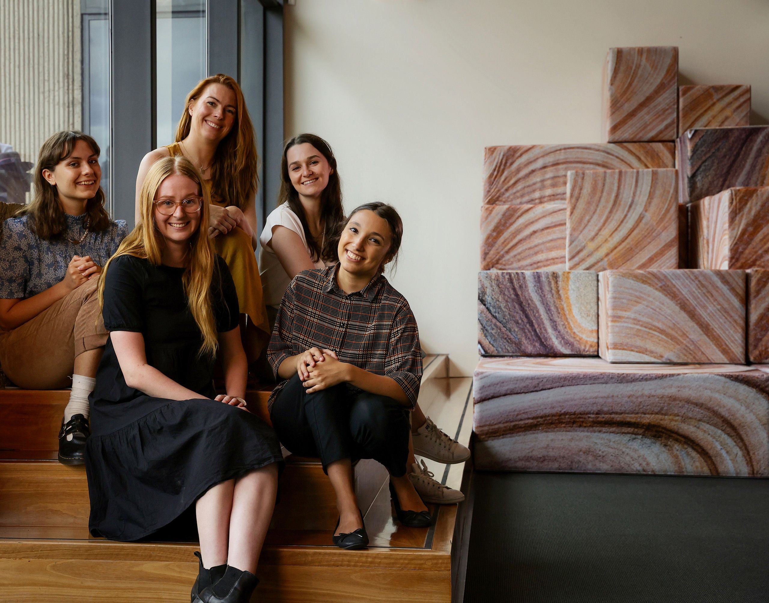 Five young women who were UQ Art Museum interns are seated smiling and chatting beside the 'DEMOS' artwork in UQ Art Museum 
