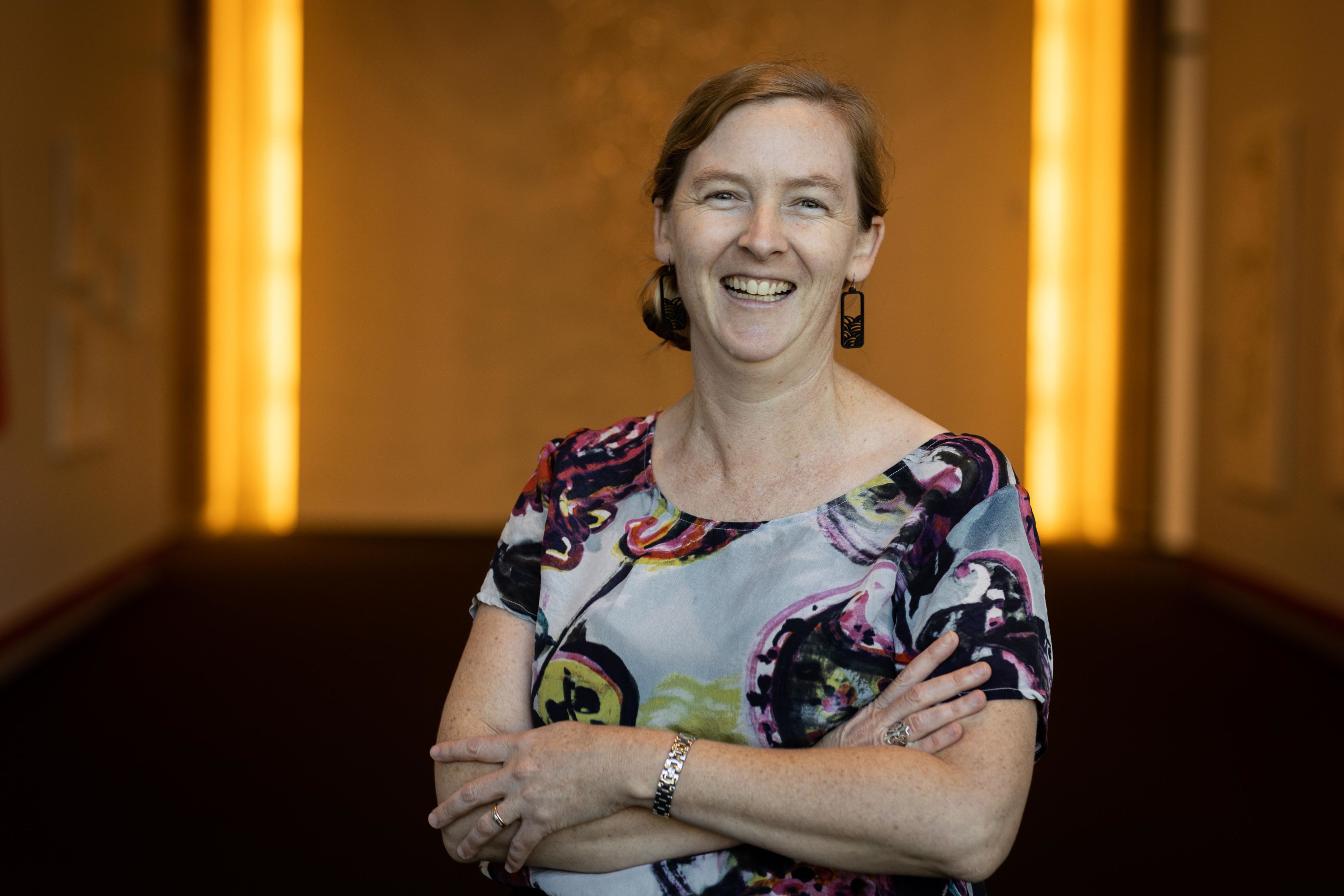 A person wwith their arms folded as they stand in front of a wall and smile at the camera