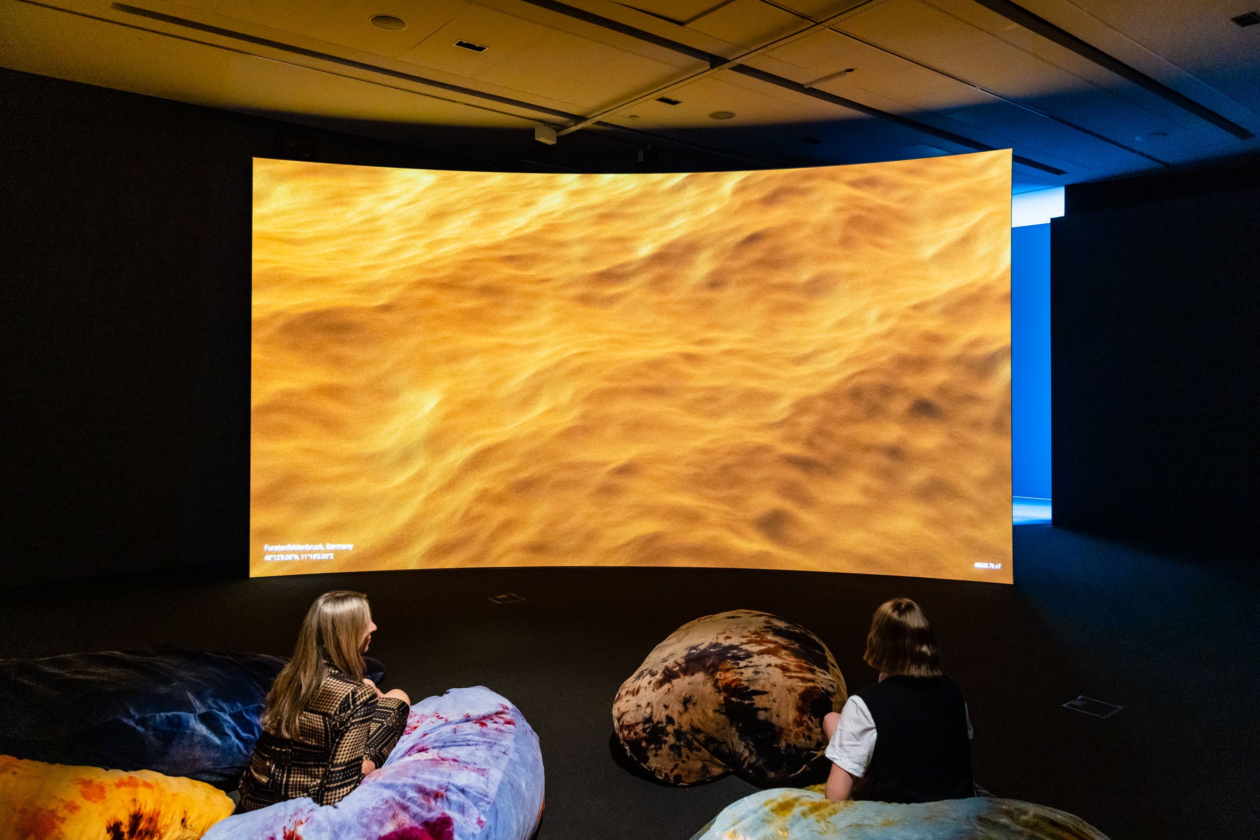Two people sitting in front of a large curved screen with an image of golden wave textures on it