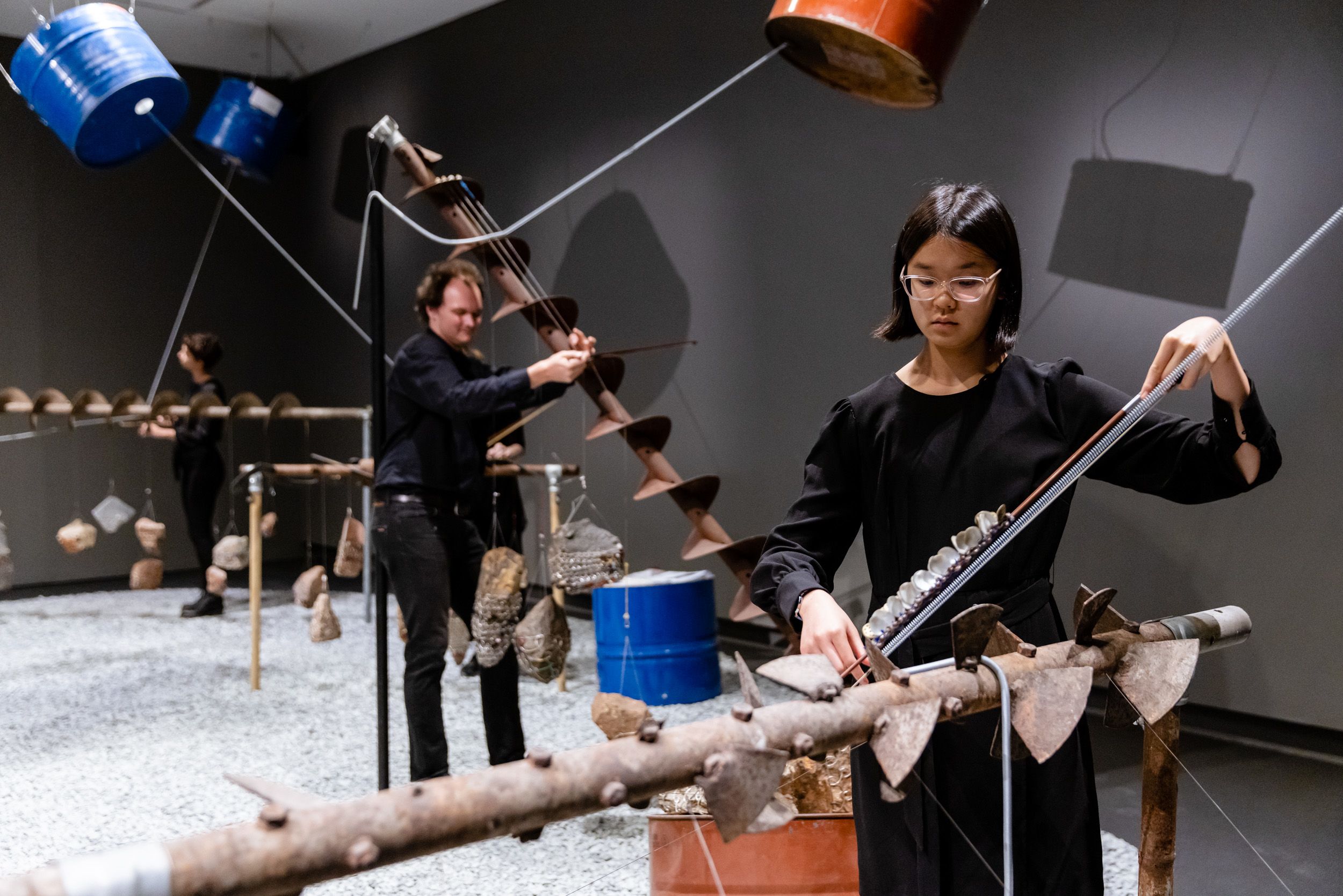 Two people playing musical instruments that resemble abstract 3D sculptures made of metal, rocks, and wires