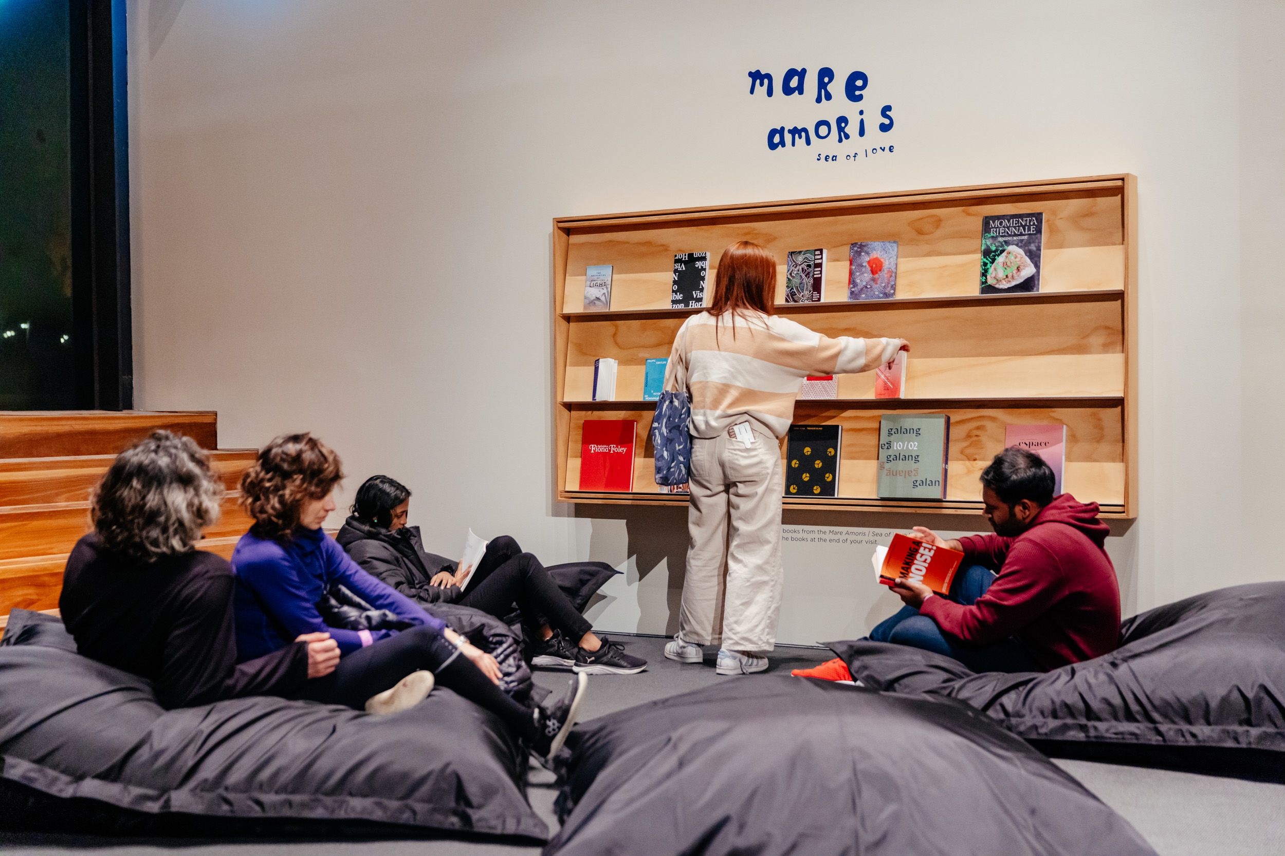 A group of people sitting on bean bags in front of a wooden library book shelf holding a collection of different coloured books