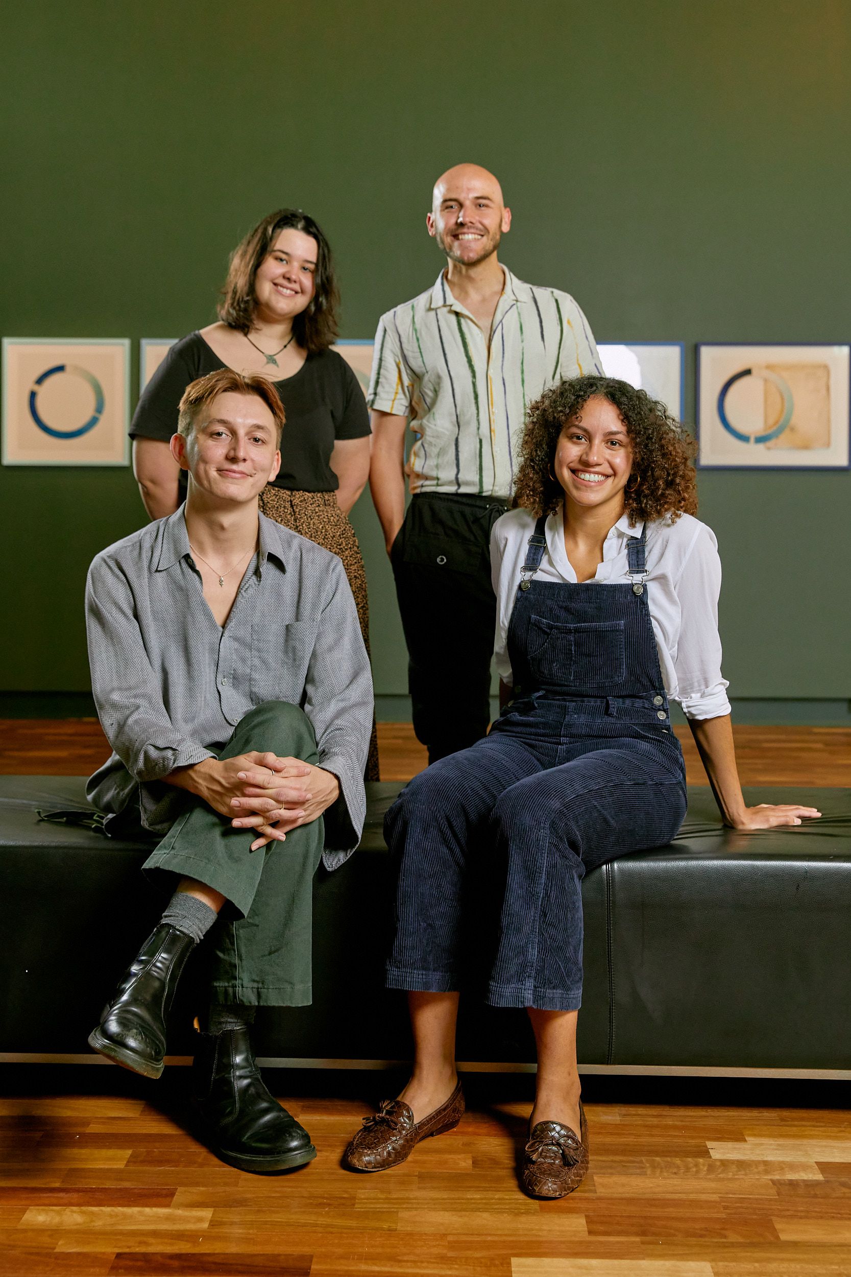 Two people sitting on a black leather ottoman with two people standing directly behind them. All are smiling.