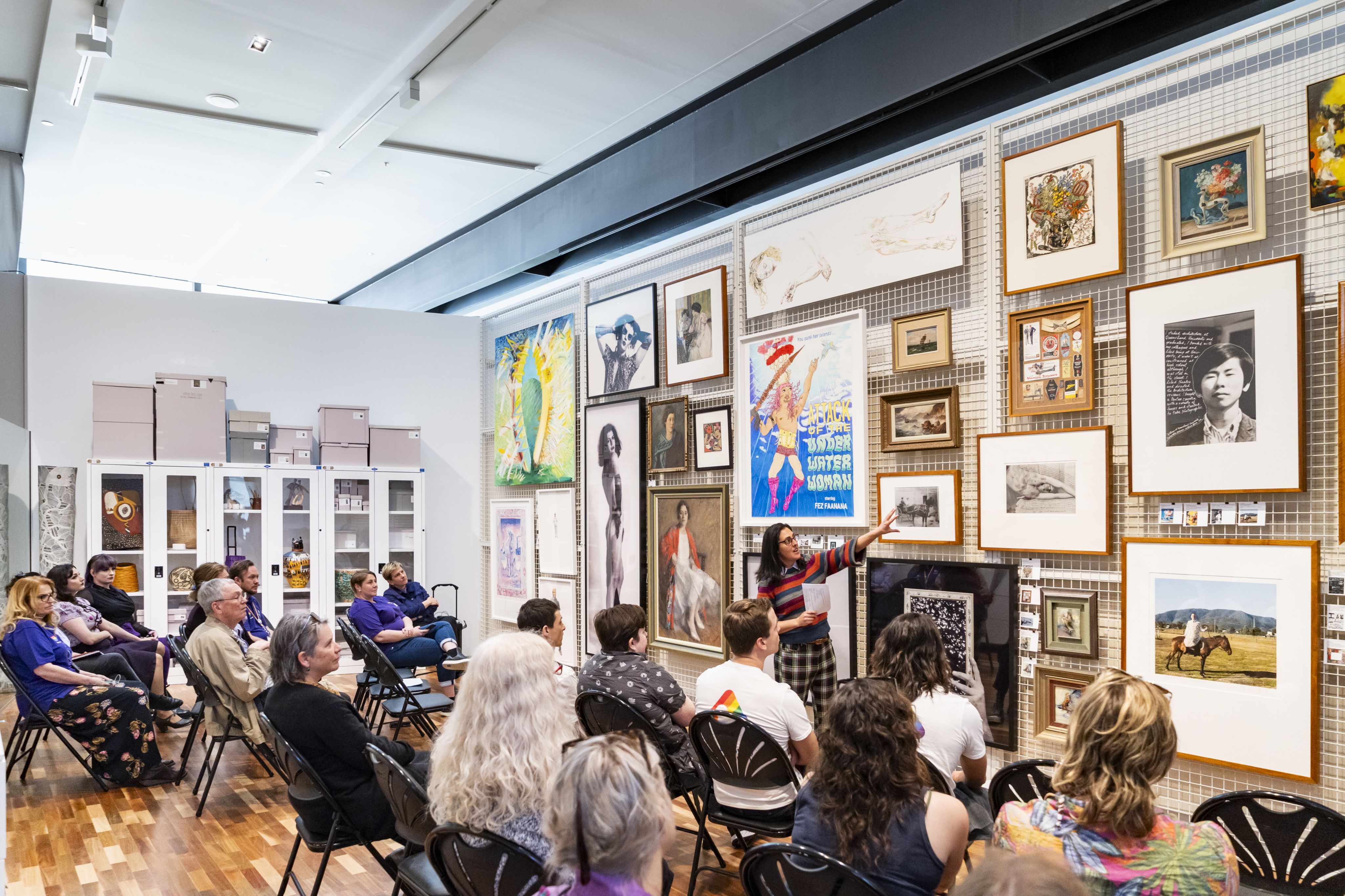 A person standing and pointing to a large wall of closely hung artwork while presenting to a group