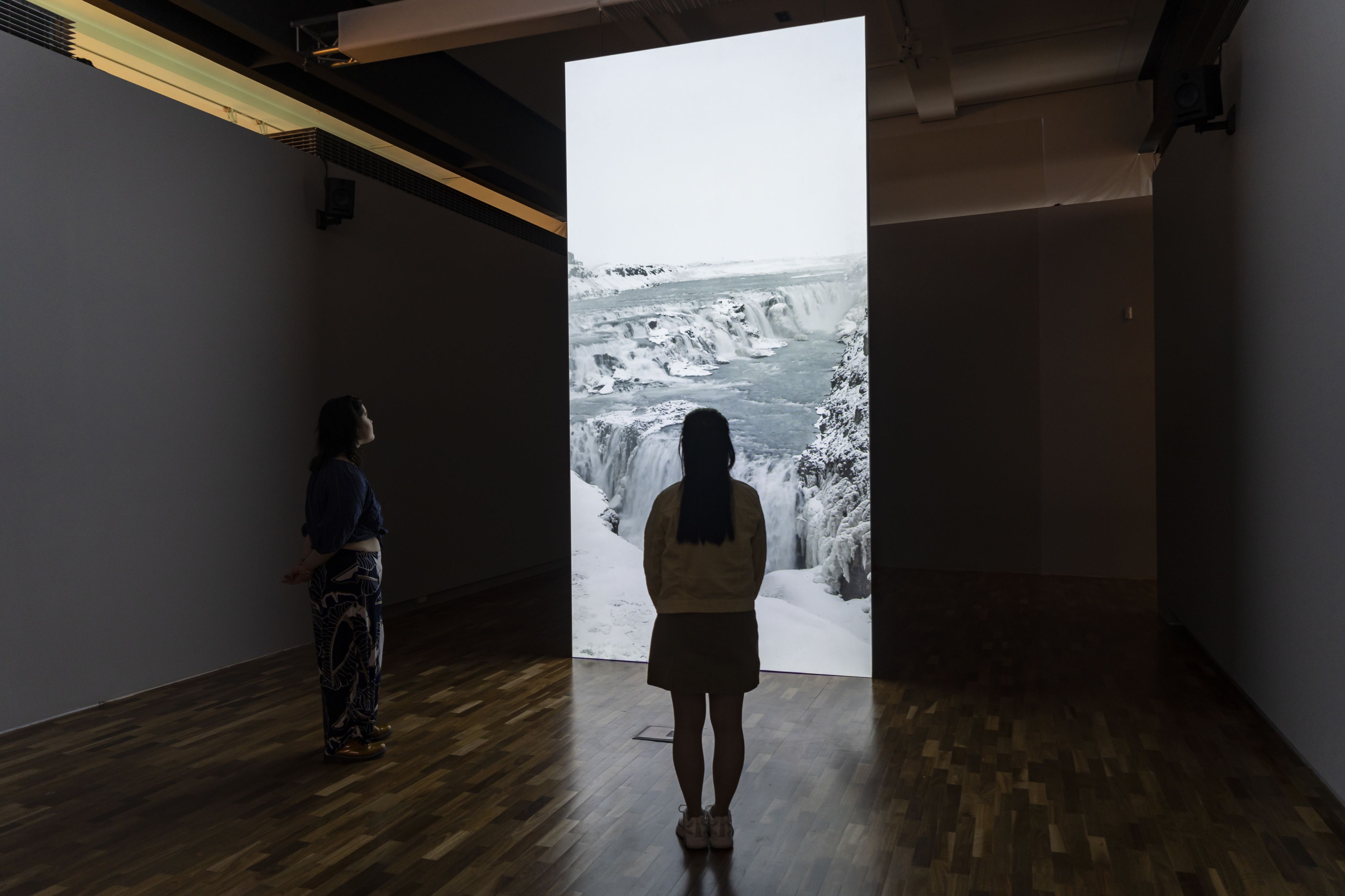 Two people standing in a dark room looking at a tall projection screen showing a snowy glacier scene.