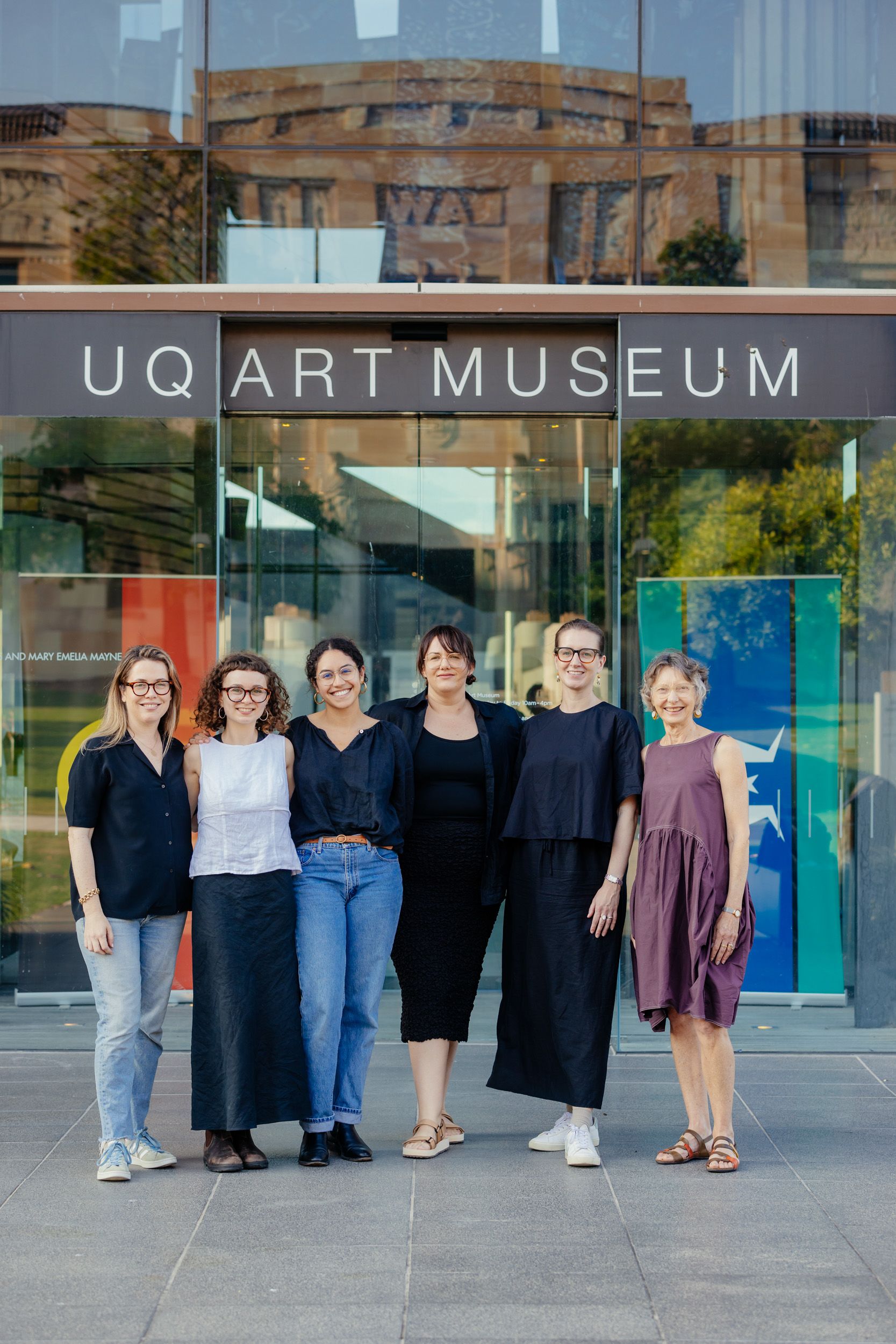 Six people stand closely together smiling at the viewer under a sign reading 'UQ Art Museum' hanging above glass sliding doors.
