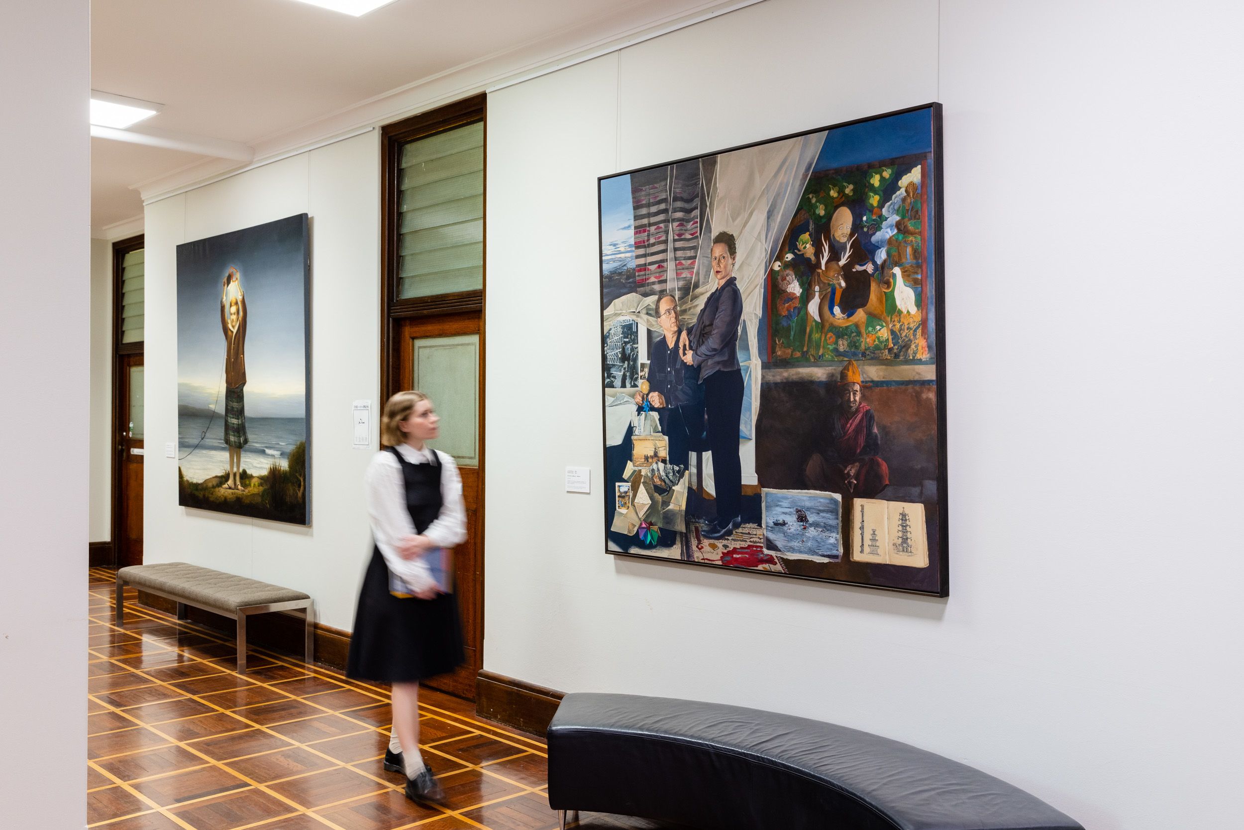A person walking past two large paintings featuring people in different surreal settings