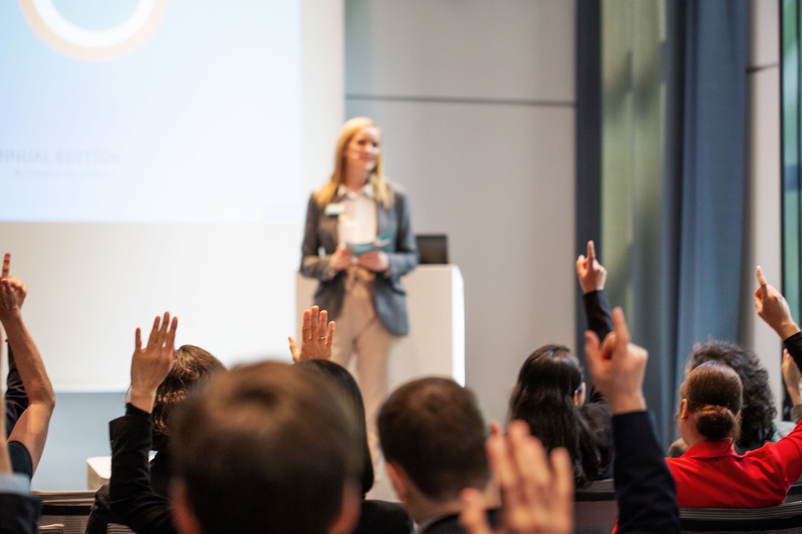 Business professional in front of crowd raising hands to ask questions.