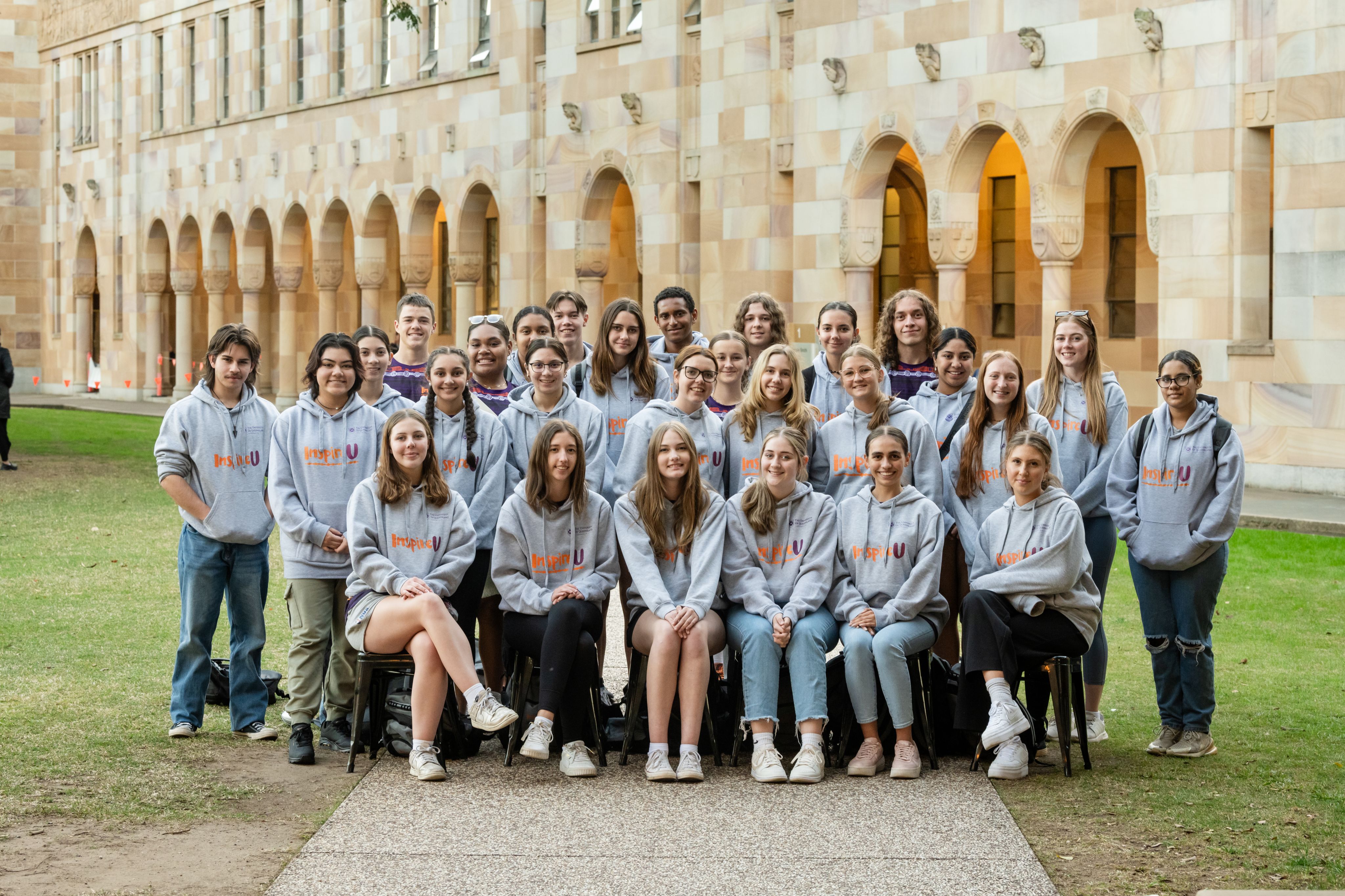A group of young people wearing grey hooded jumpers, standing on the grassy lawn in front of a sandstone building