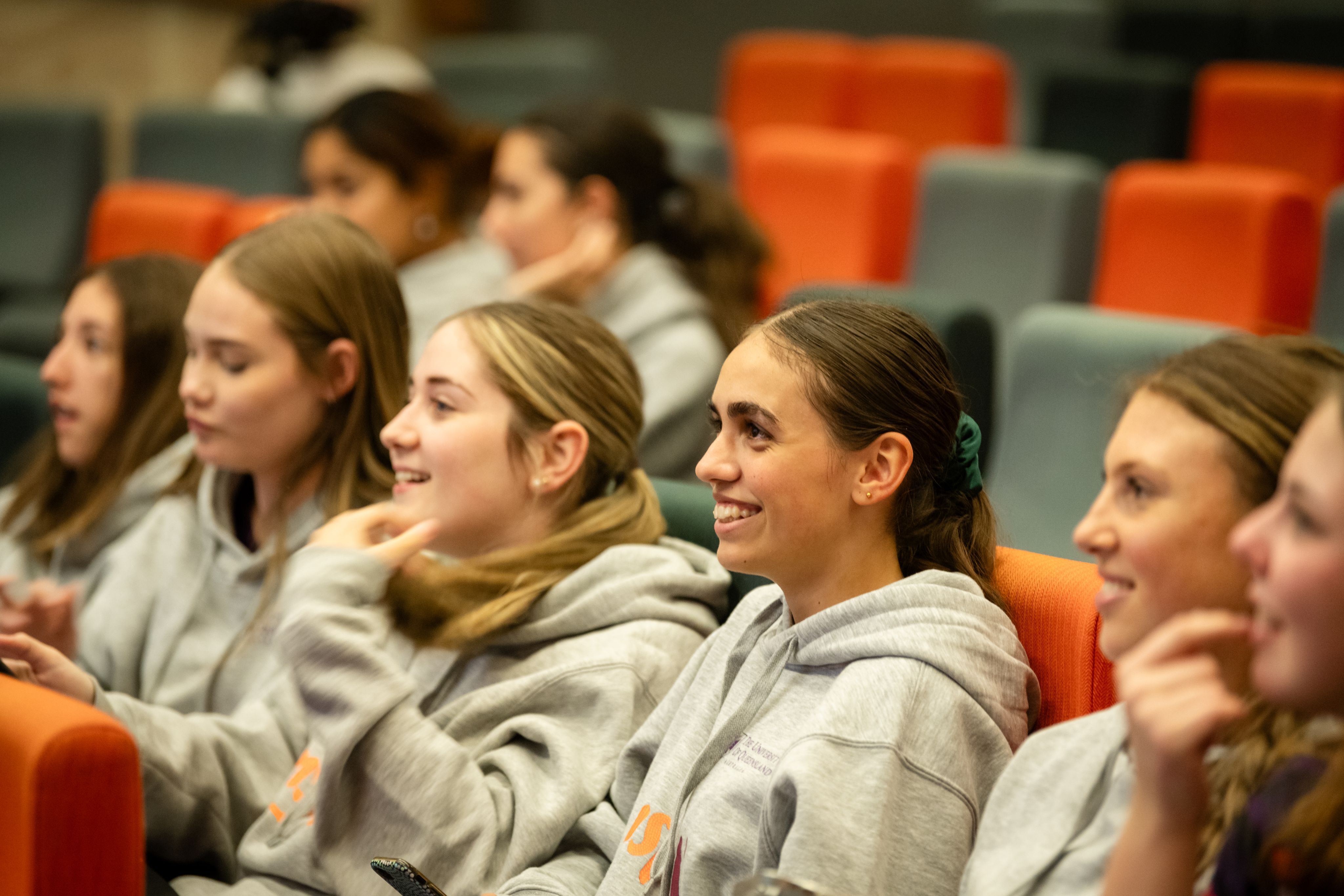Young women wearing grey hooded jumpers, sitting in grey and orange theatre chairs and smiling
