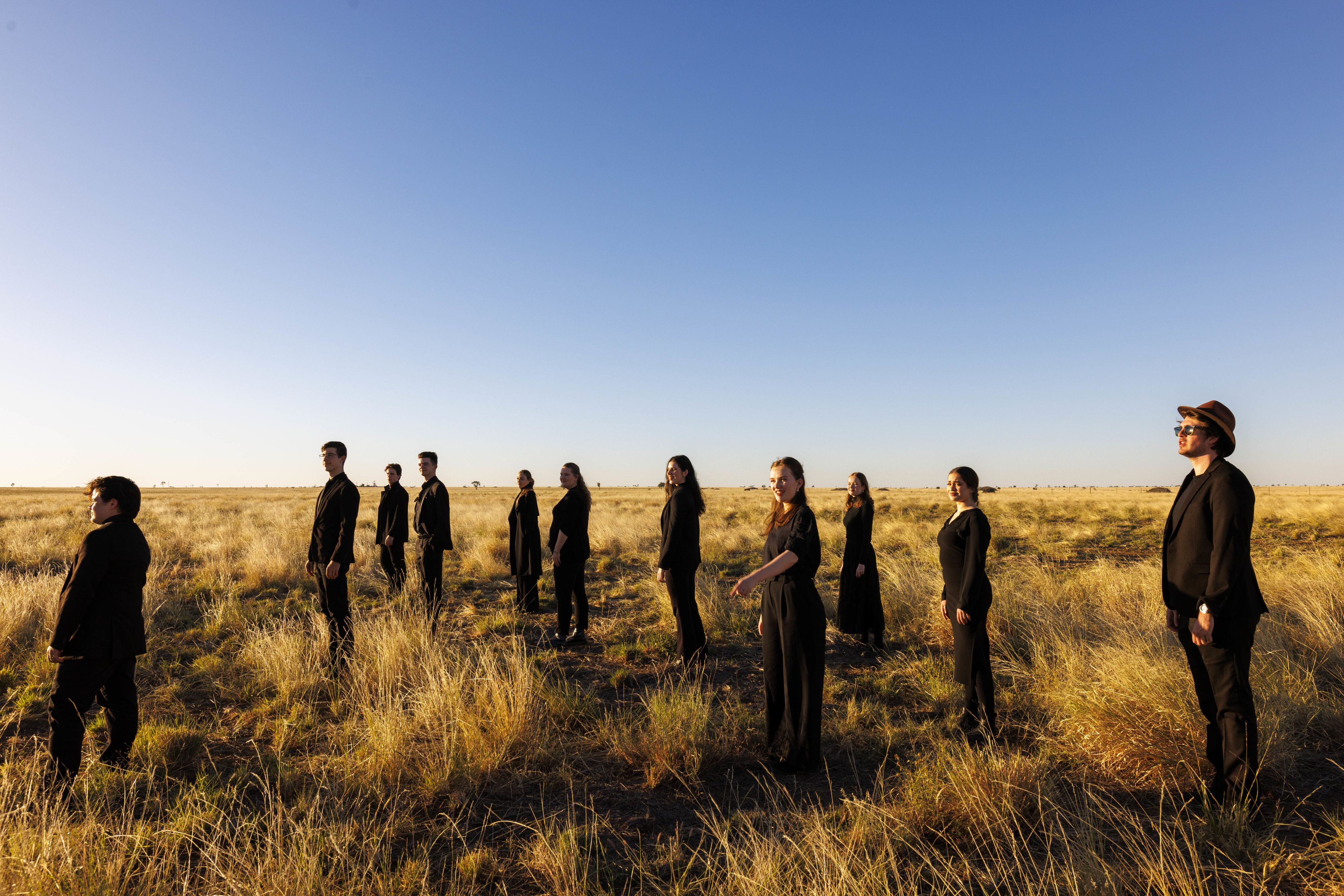 11 students in black outfits stand in a dry grass field under a blue sky as the sun sets