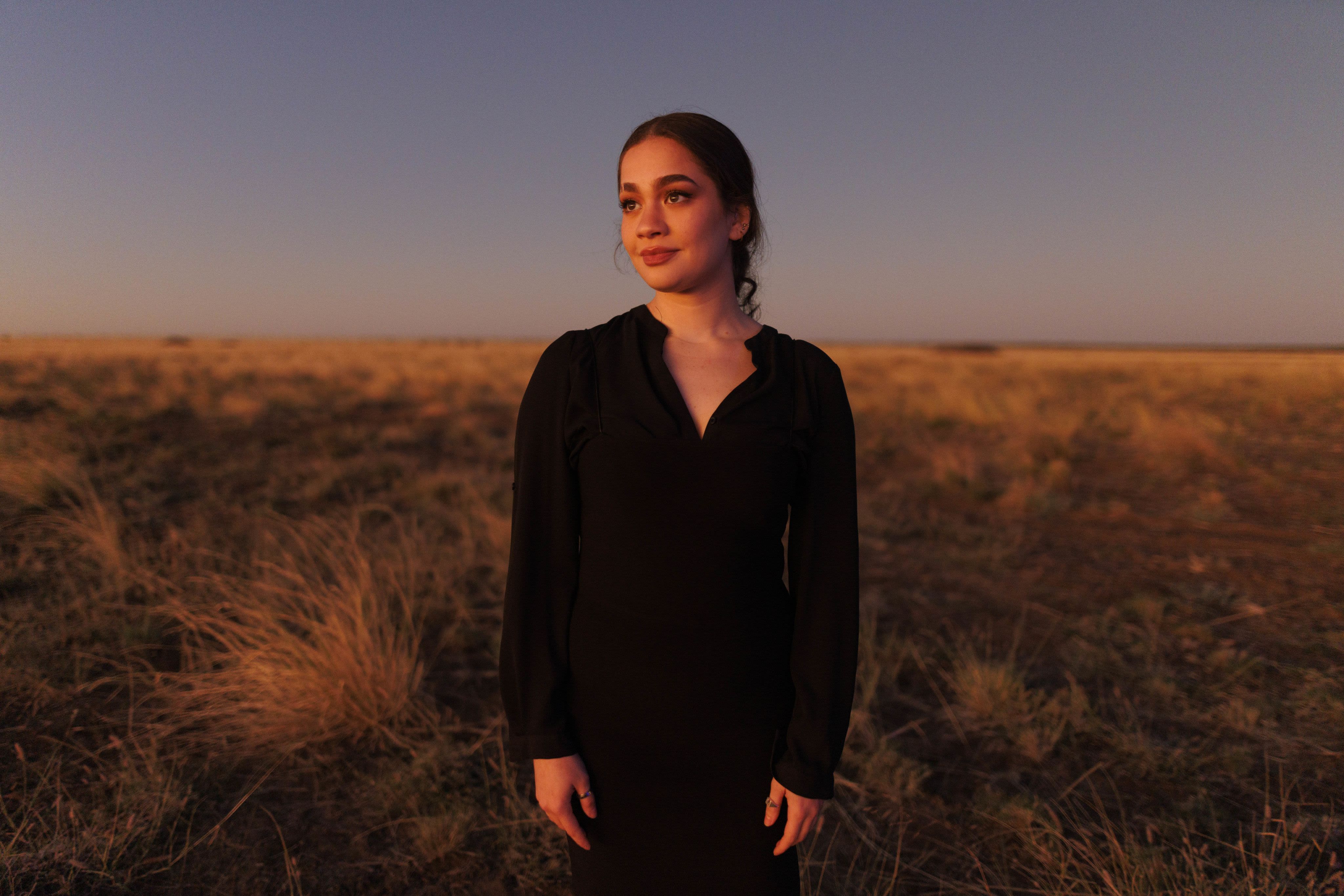A young woman wearing a black t-shirt standing in a grassy field under the warm light of sunset