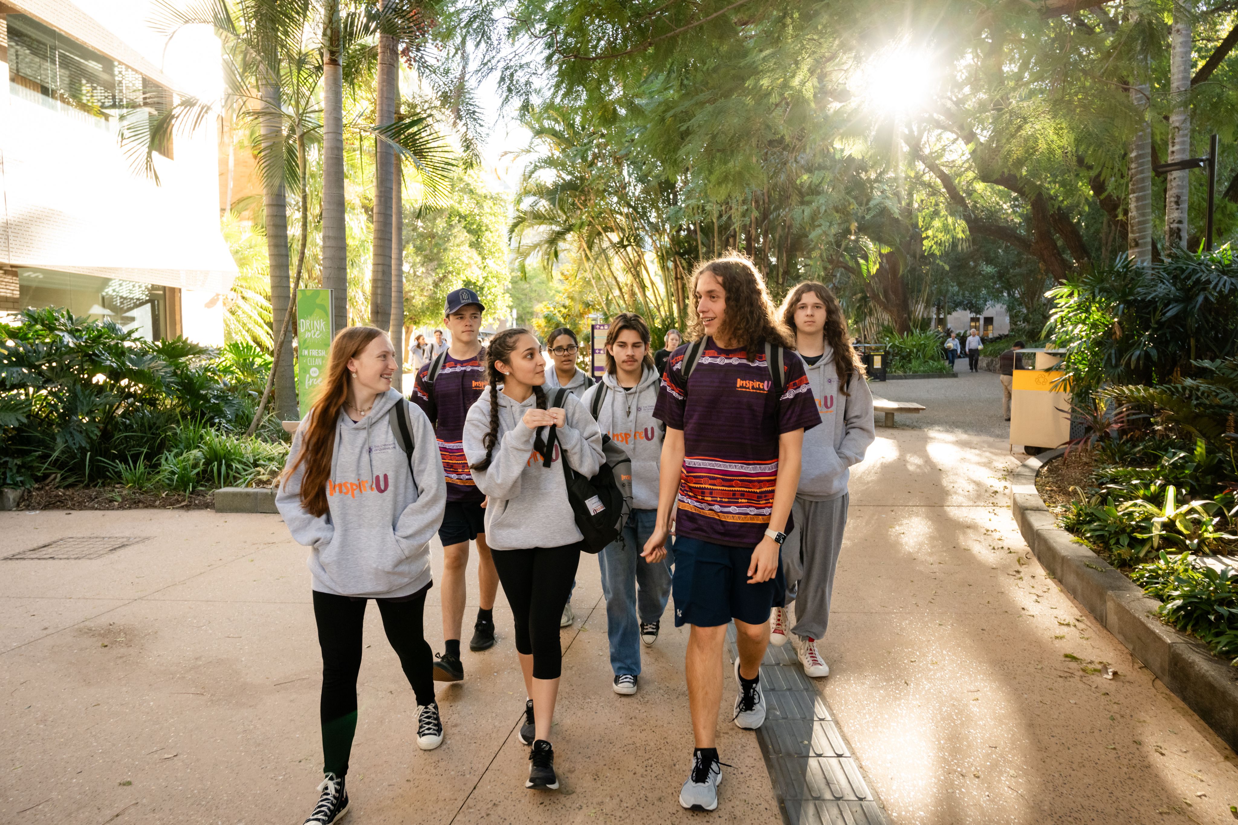 A group of young people wearing grey jumpers, with two people wearing purple and orange patterned t-shirts. They are walking through a lush rainforest area of a university campus