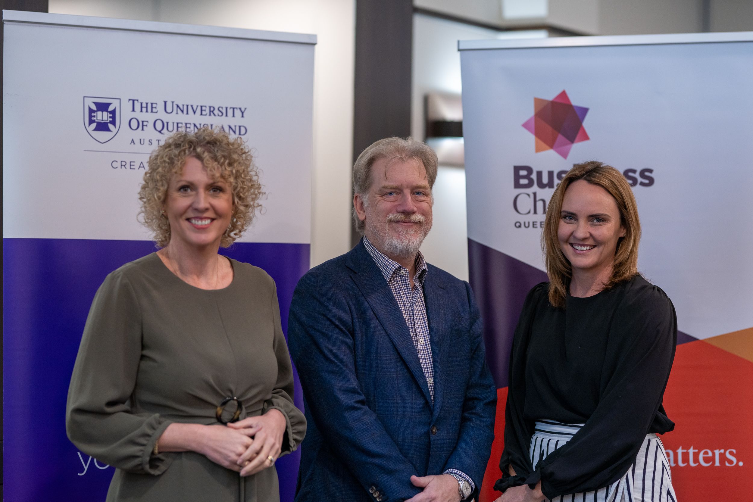 Two women and one man standing in front of a University of Queensland banner and a Business Chamber Queensland banner
