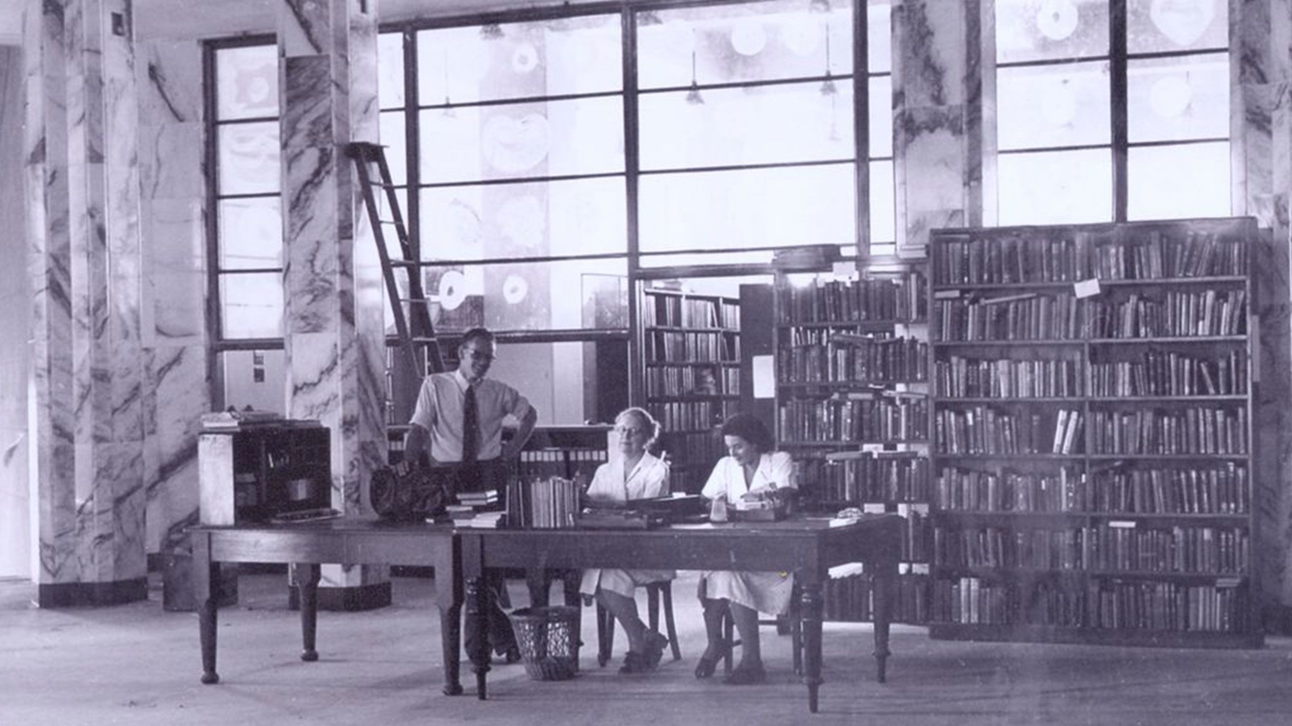 An image of library staff, including Harrison Bryan and Nellie McIver, at the desk in the Main Library in 1949.