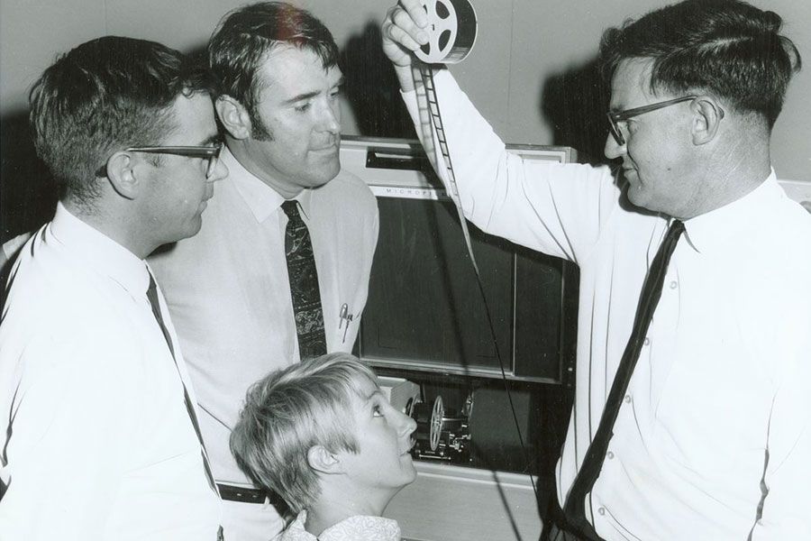 An image of Spencer Routh, Frank Thompson (Manager of UQ Press), Sue Pechey and Roger Joyce examine historical microfilm of Pugh's Almanac in the Main Library in 1968.