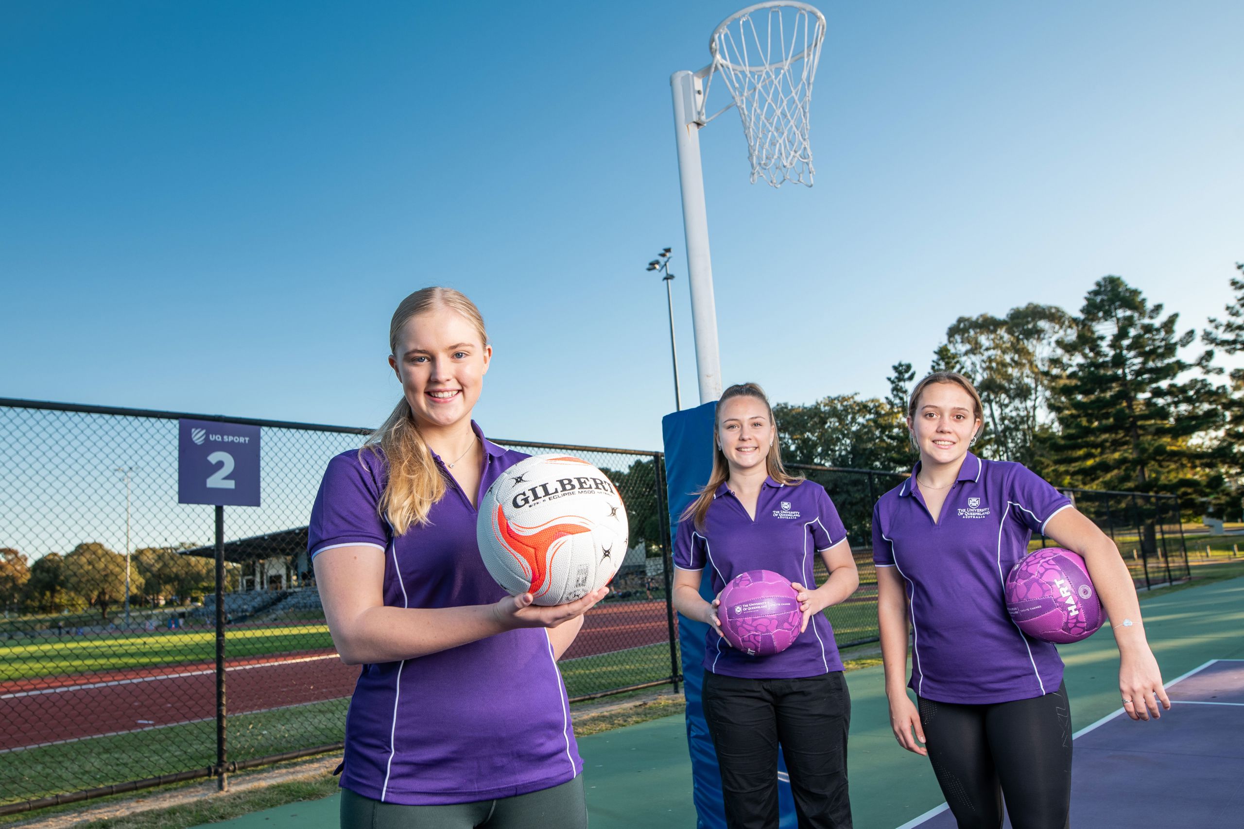 An image of UQ Firebirds Scholarship recipients Savanna Detlefson, Brooke Warburton, and Alyssa Hanson holding netbals at the netball courts at UQ's St Lucia campus.
