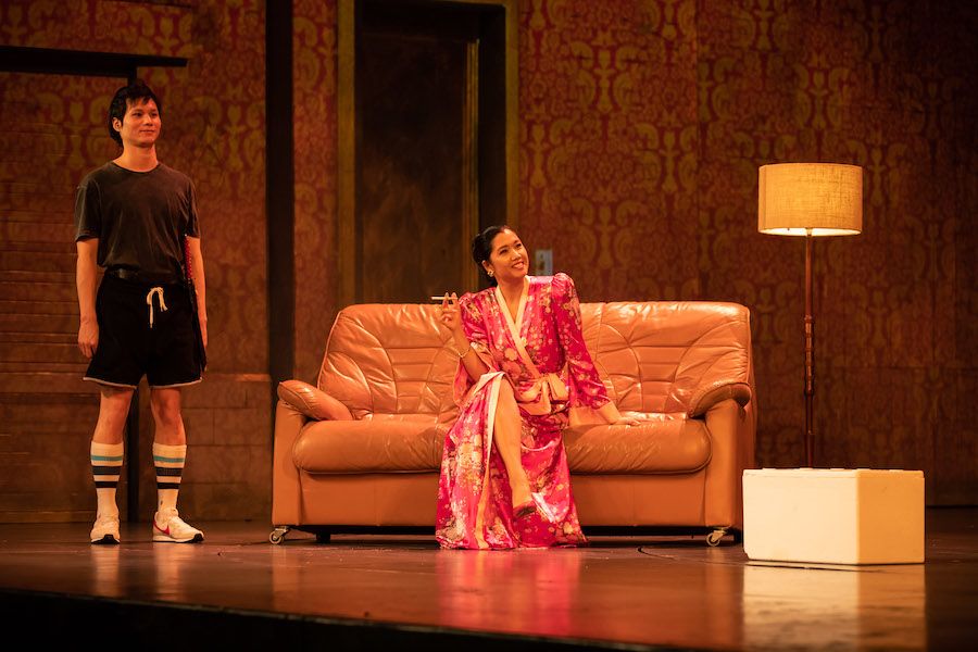 A woman sits onstage on a couch, wearing a pink silk robe and a cigarette. She is smiling. Next to her stands a young man in a black t-shirt, gym shorts, tall white socks and joggers.