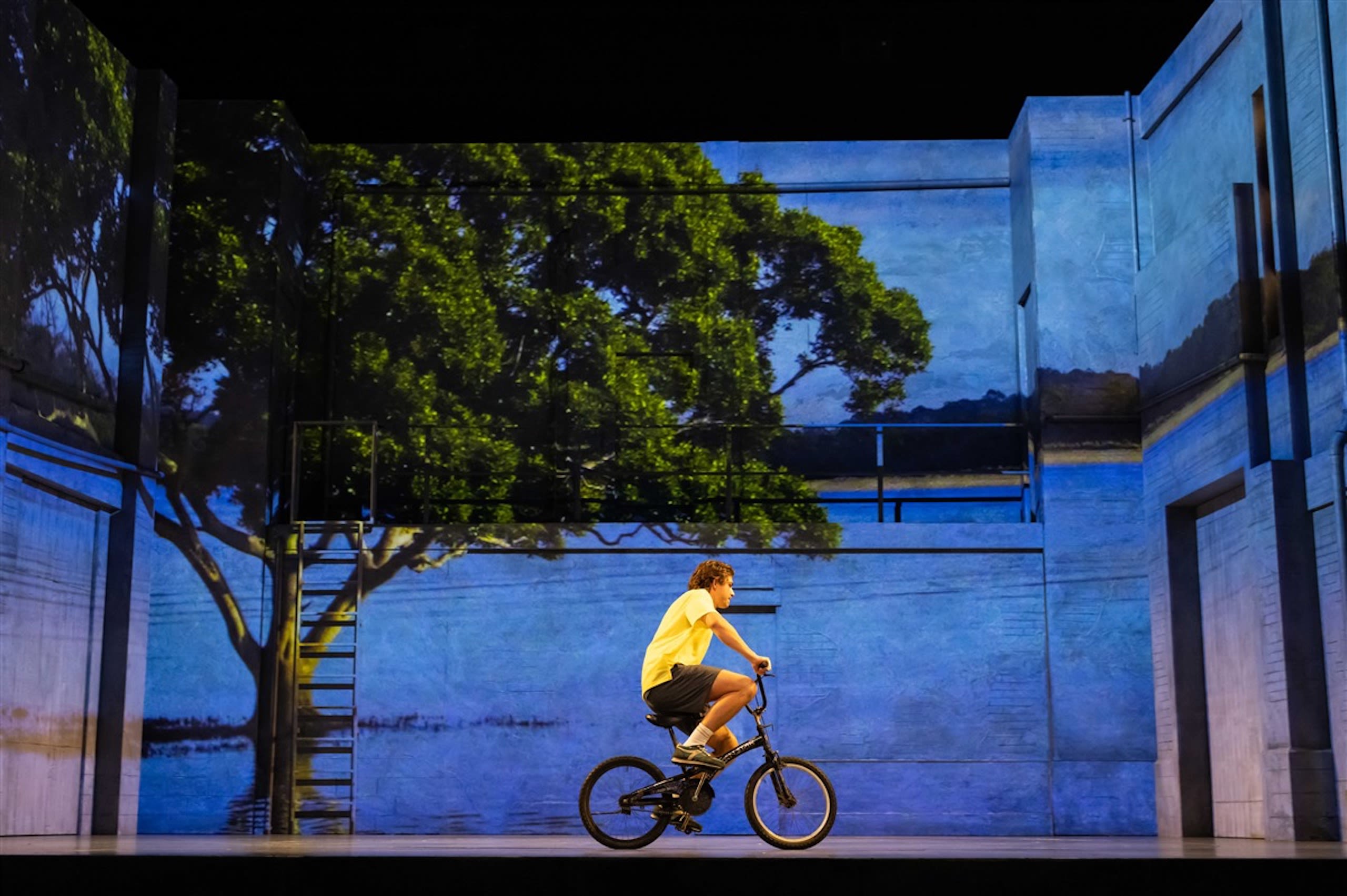 A young man wearing a yellow t-shirt rides a bicycle on a theatre stage.