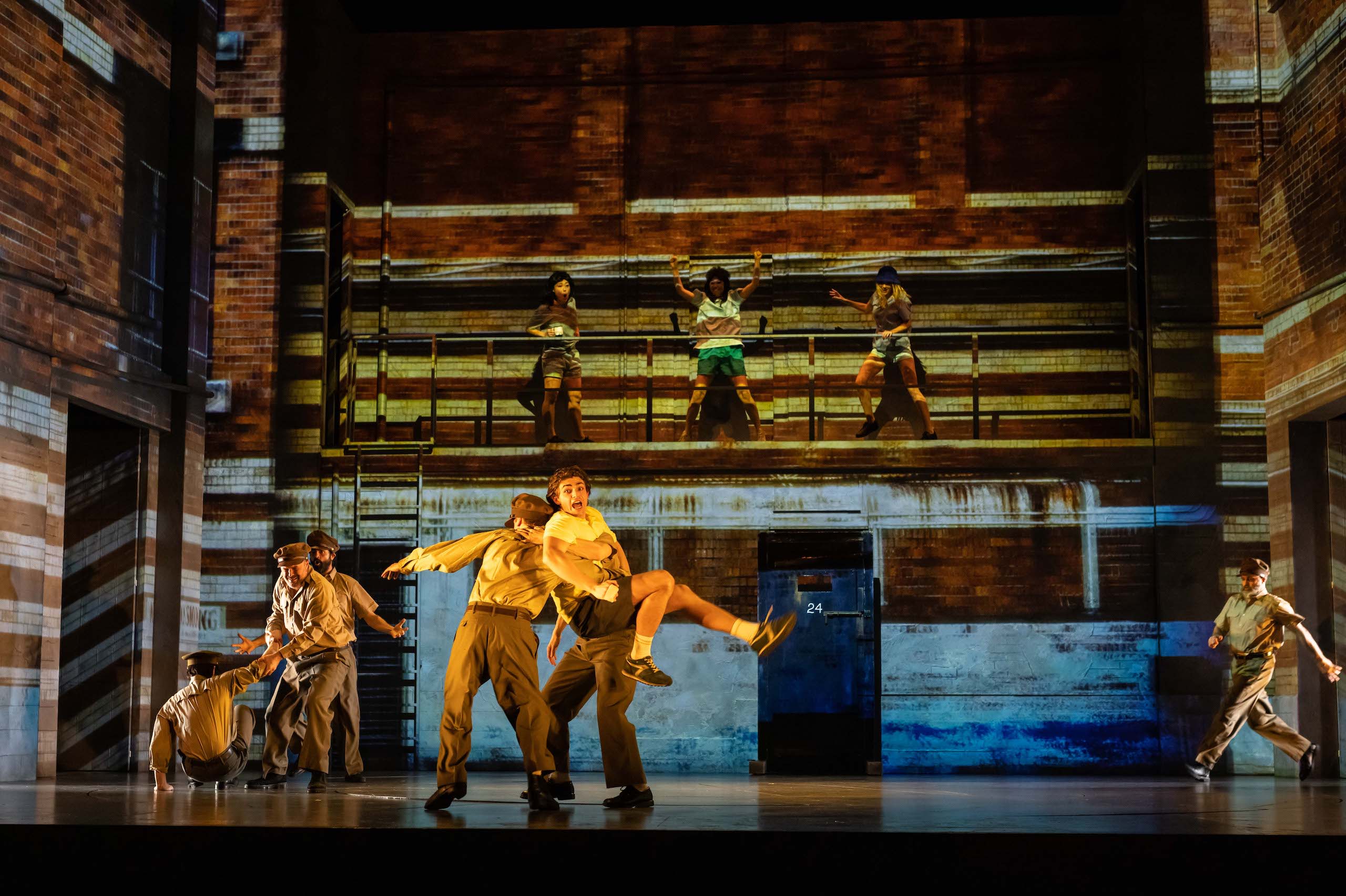 A stage scene where a young man is being carried by two security guards in a jail setting.