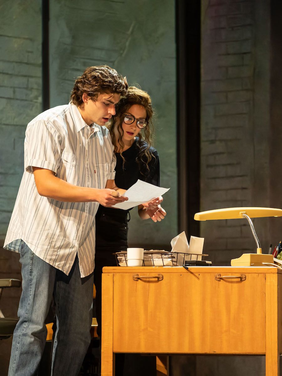 A young man and a young woman stand onstage lit by a bright light. They are huddled together reading a letter.