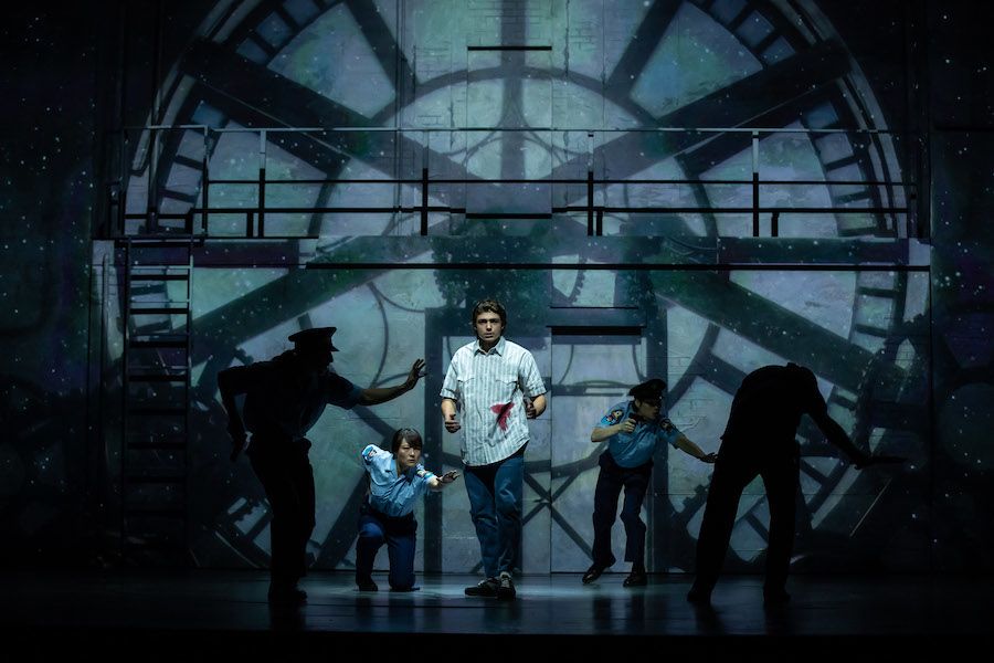 A young man in a blue and white striped button down stands in the middle of a stage. In the background is the silhouette of a large clockface, depicting the inside of a clock tower.