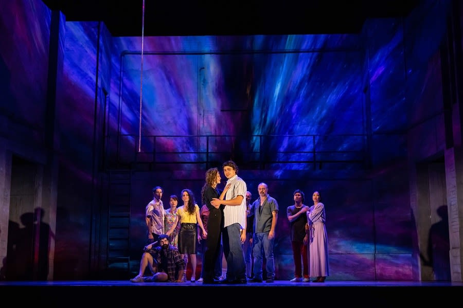 Seven actors stand onstage facing the audience. The stage is lit with blue and purple lights.