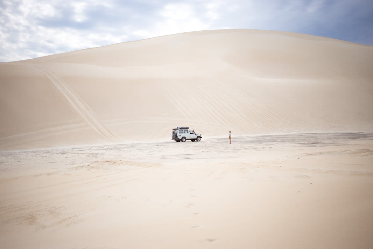 A white Toyota Landcruiser parked in front of a towering beige sand dune.