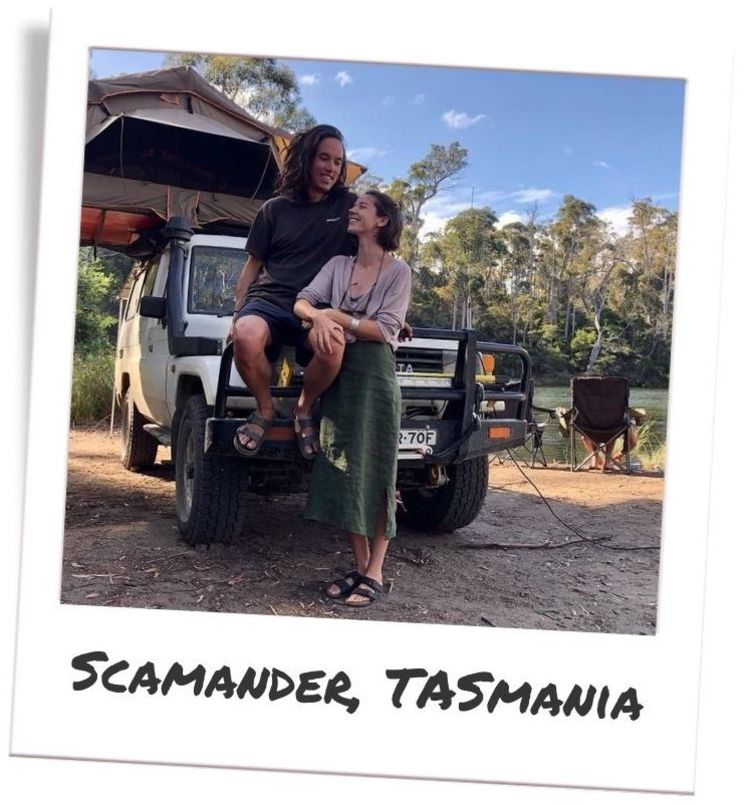 A young adult couple smiling at each other in front of their white Toyota Landcruiser troop carrier vehicle. Behind them are camp chairs by a stream.