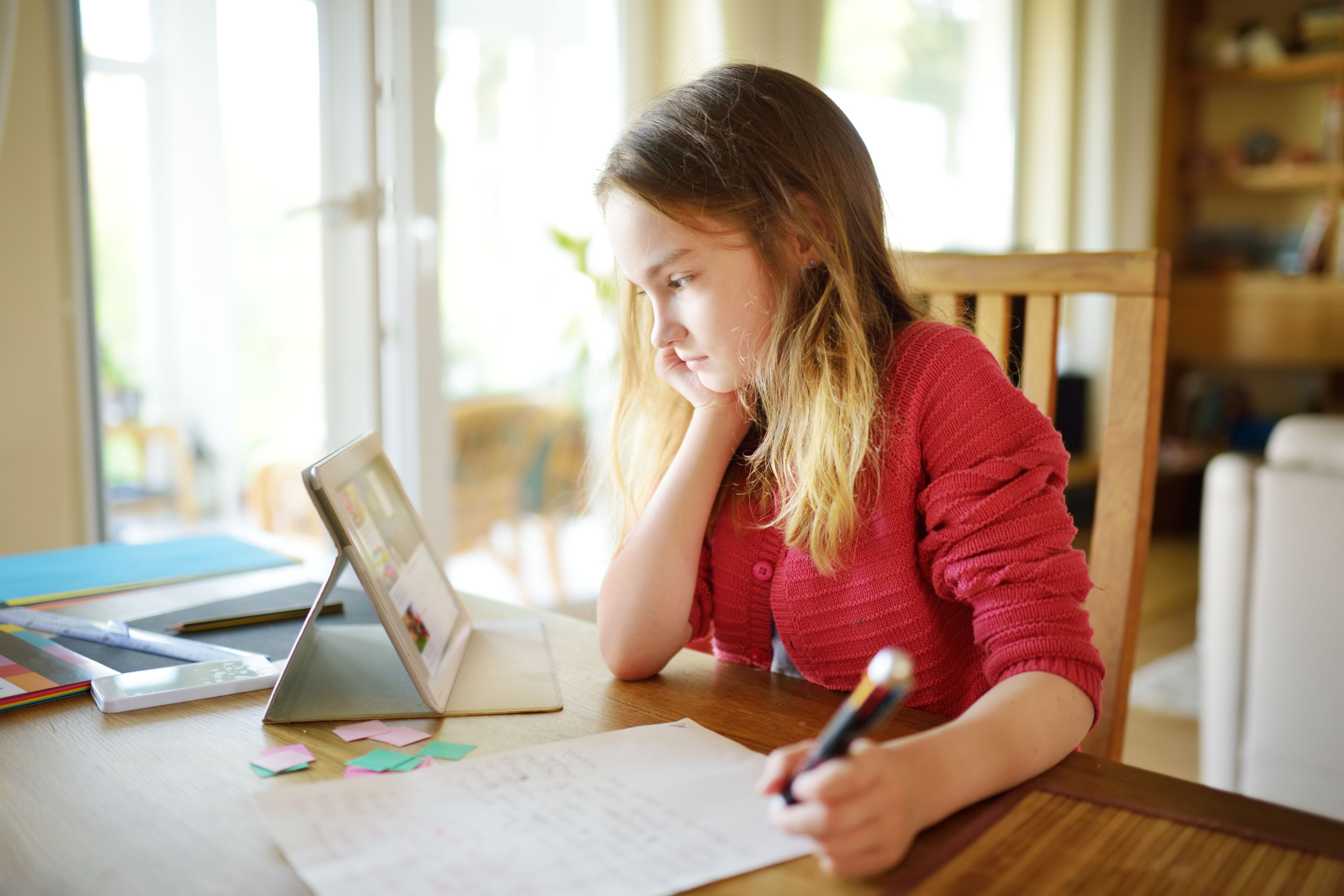 Young girl writing on a piece of paper in front of a tablet.